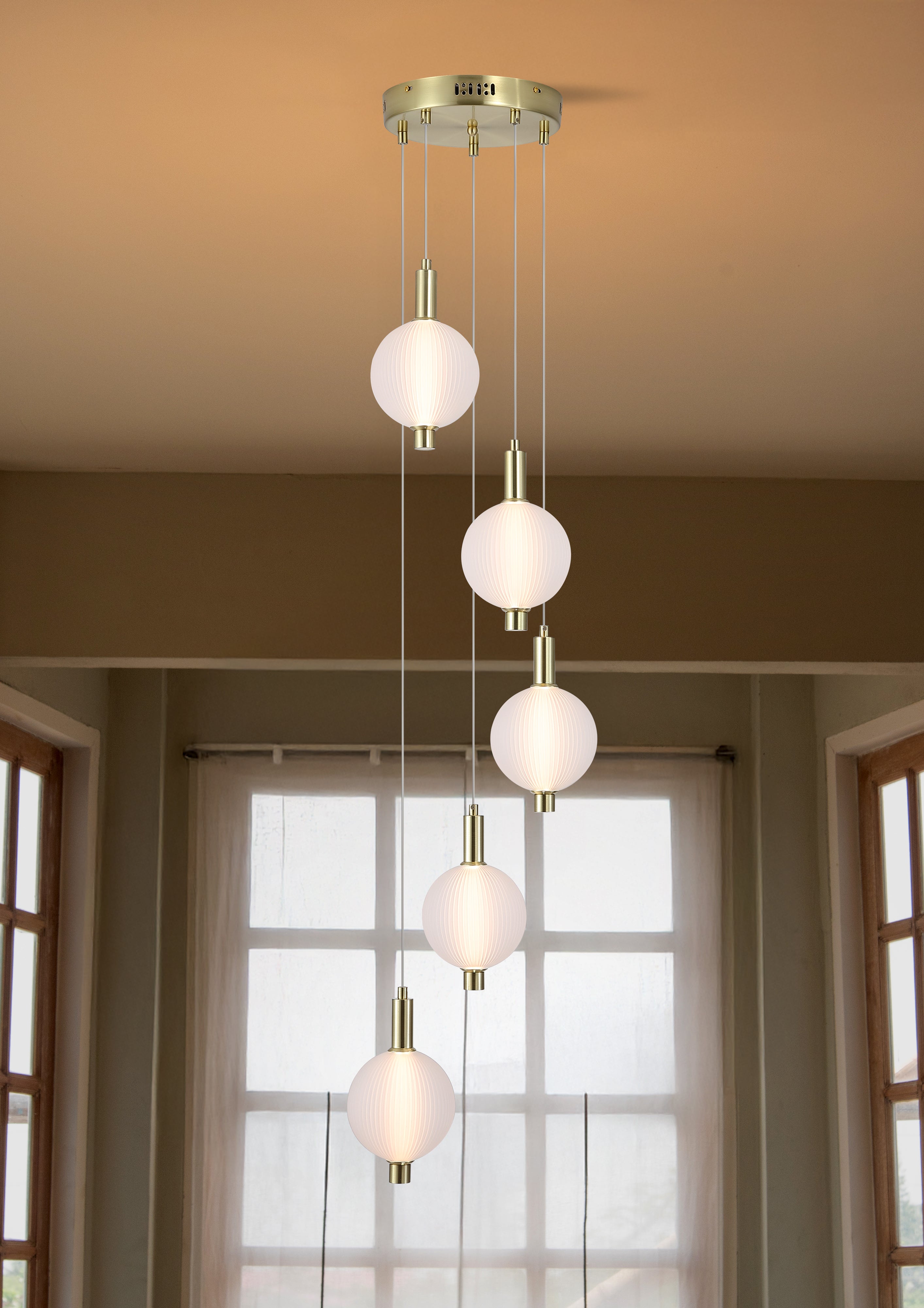 Searchlight Luminique LED 5 light pendant in satin brass, hanging lit with staggered globes in a warm toned room with tall windows