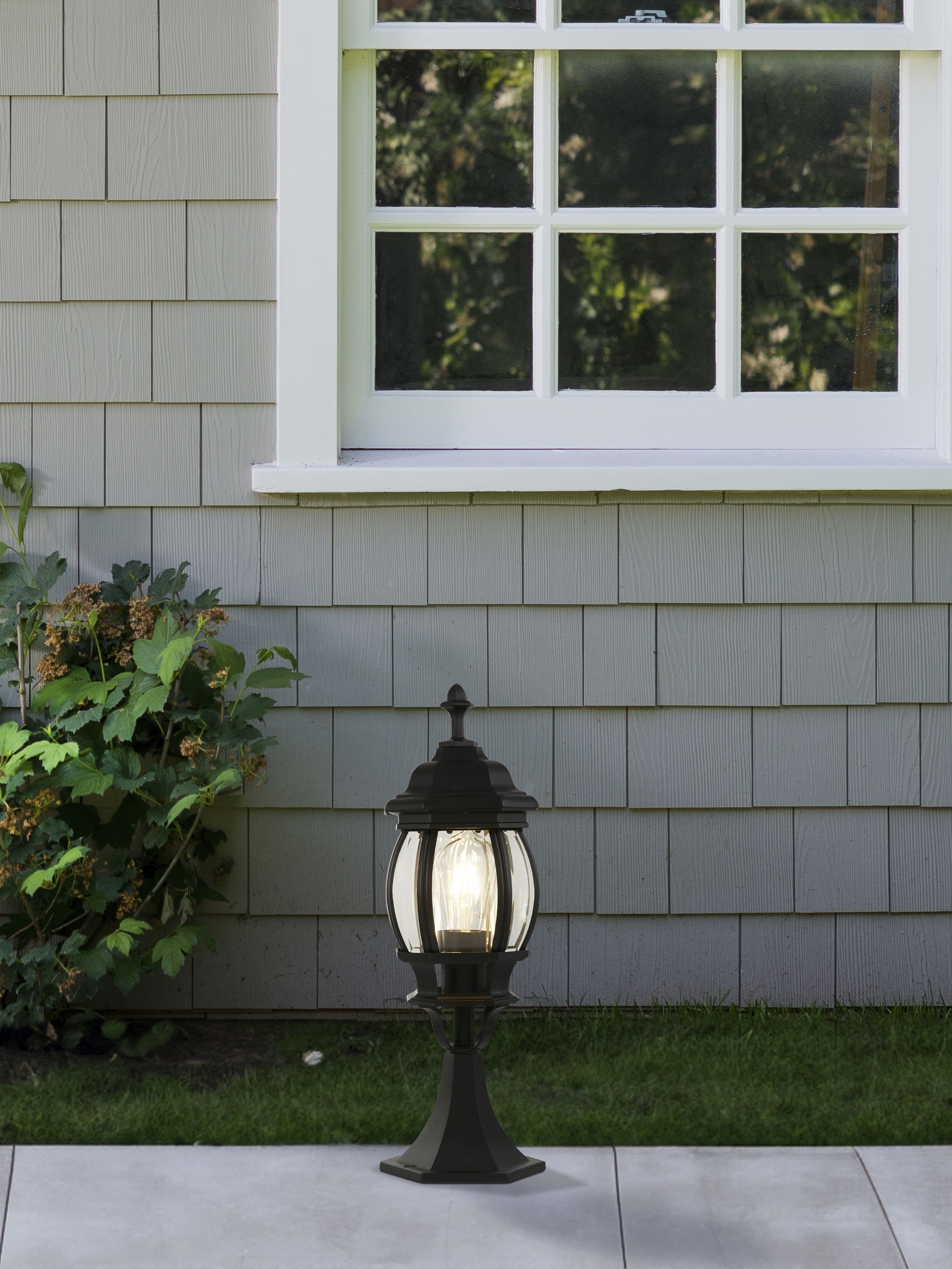 Bel Aire Outdoor Post 500mm lit beside a house wall under a window, with green shrubs nearby