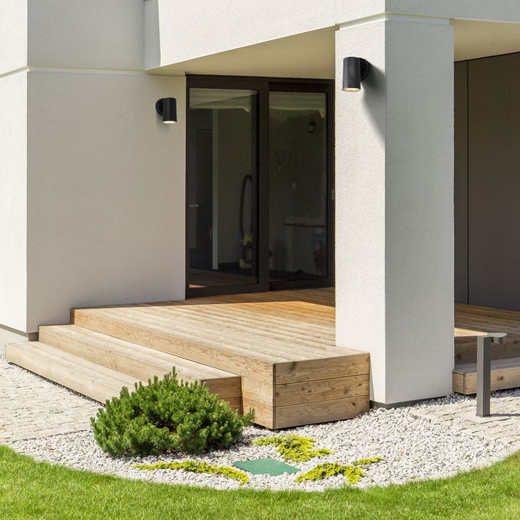 Coastal Outdoor Wall Light installed on a house exterior beside a wooden deck and gravel garden in daylight.