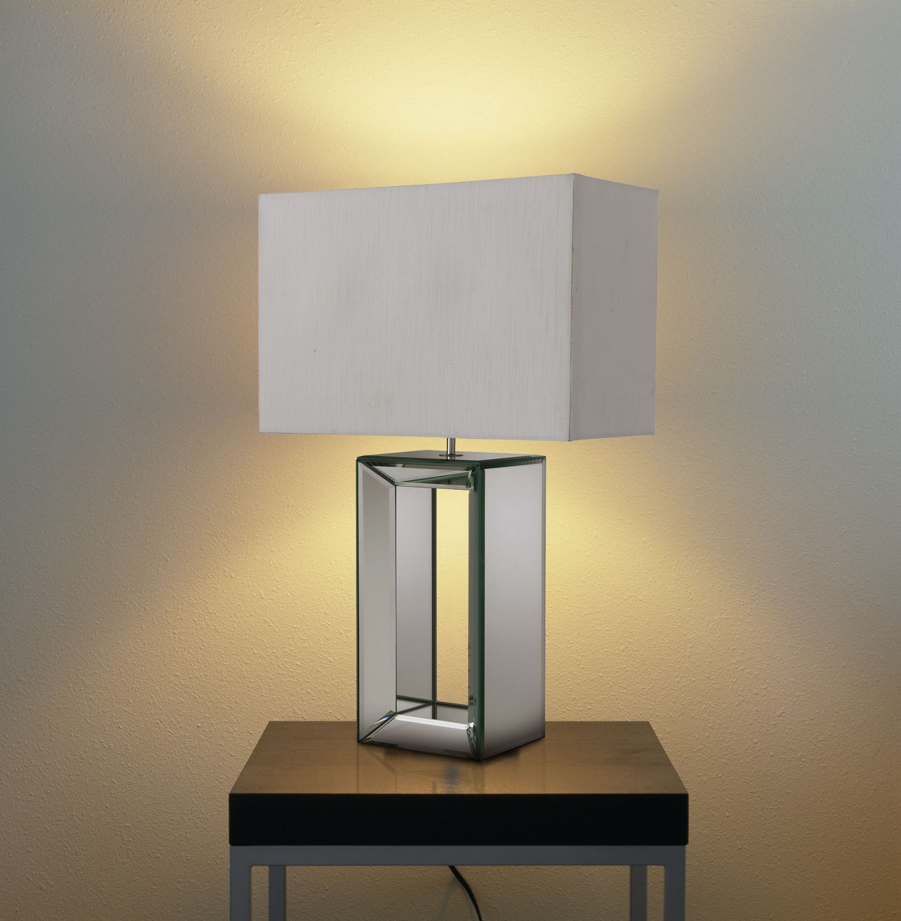 Searchlight mirror table lamp lit and styled on a dark bedside table against a warmly lit wall