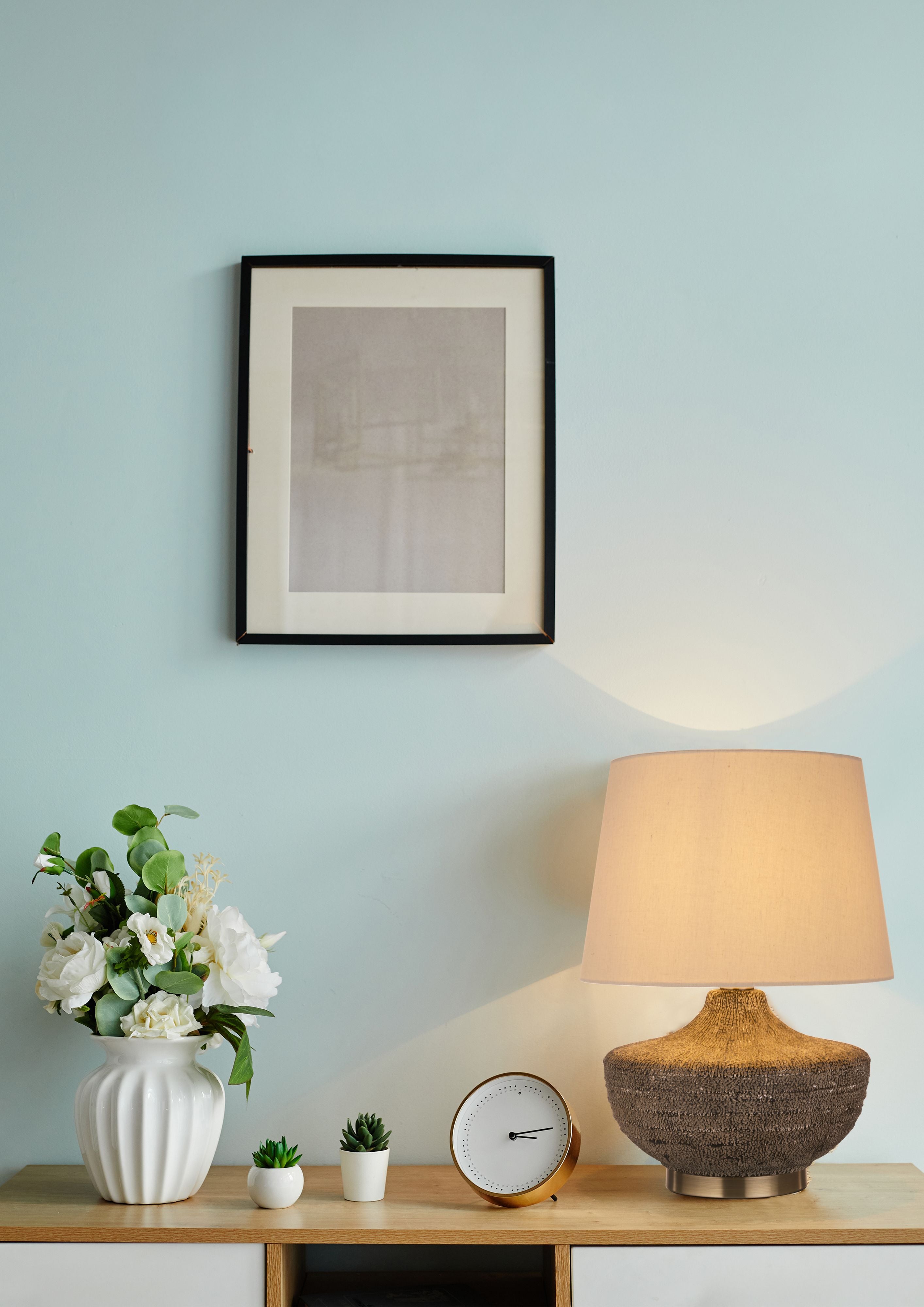 Searchlight Avaya table lamp lit on a wooden console table against a duck egg blue wall, styled with a framed print, a white vase of flowers and a small clock