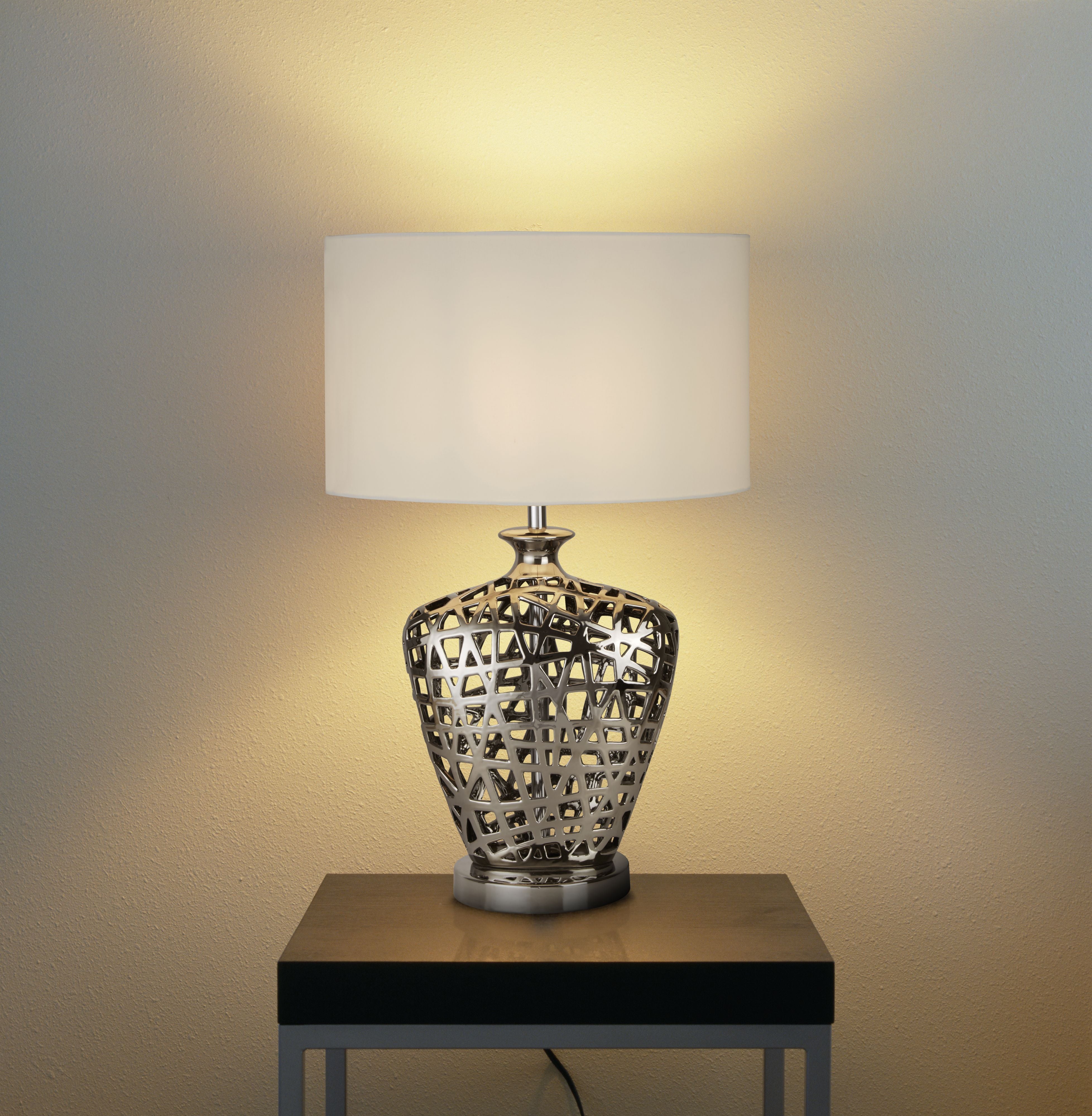 Large Network table lamp switched on and glowing warmly on a wooden side table against a plain wall