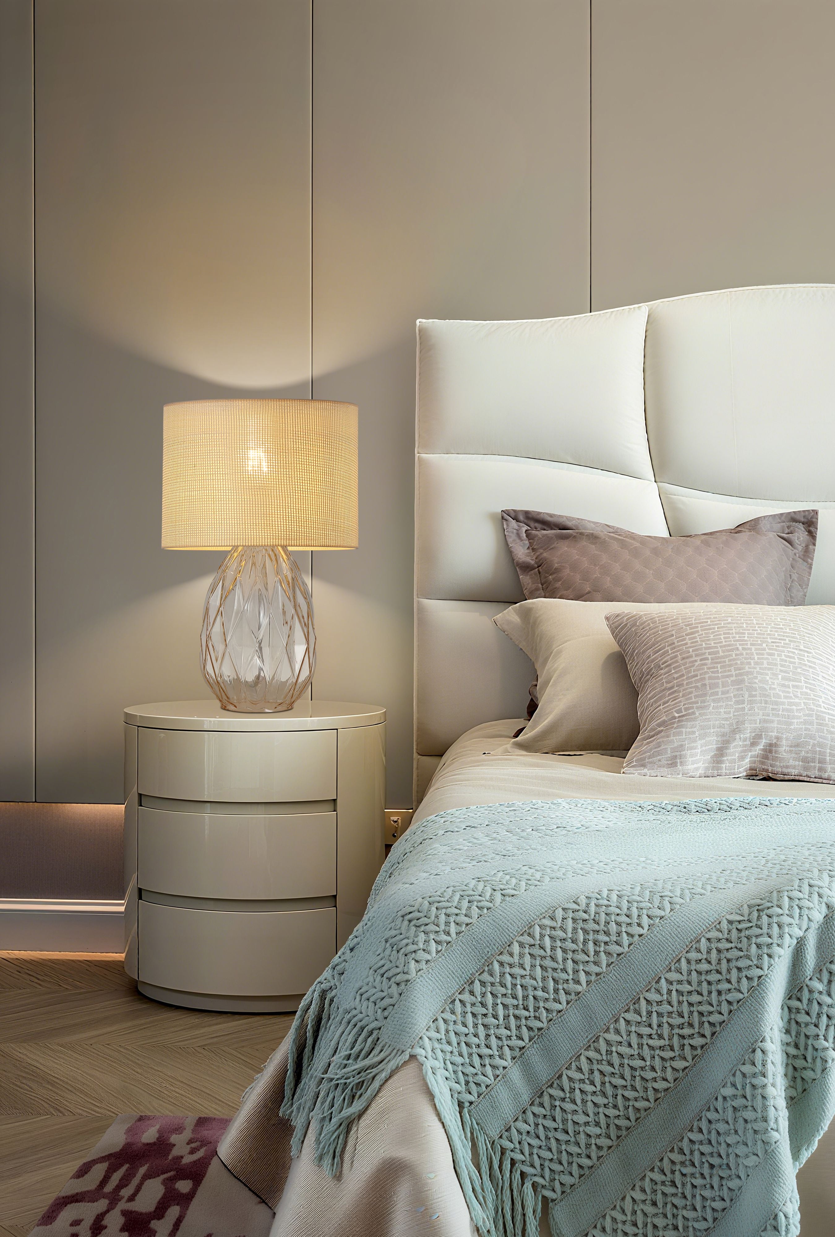 Diane table lamp lit and styled on a bedside cabinet next to a bed with pale blue throw, bedroom setting