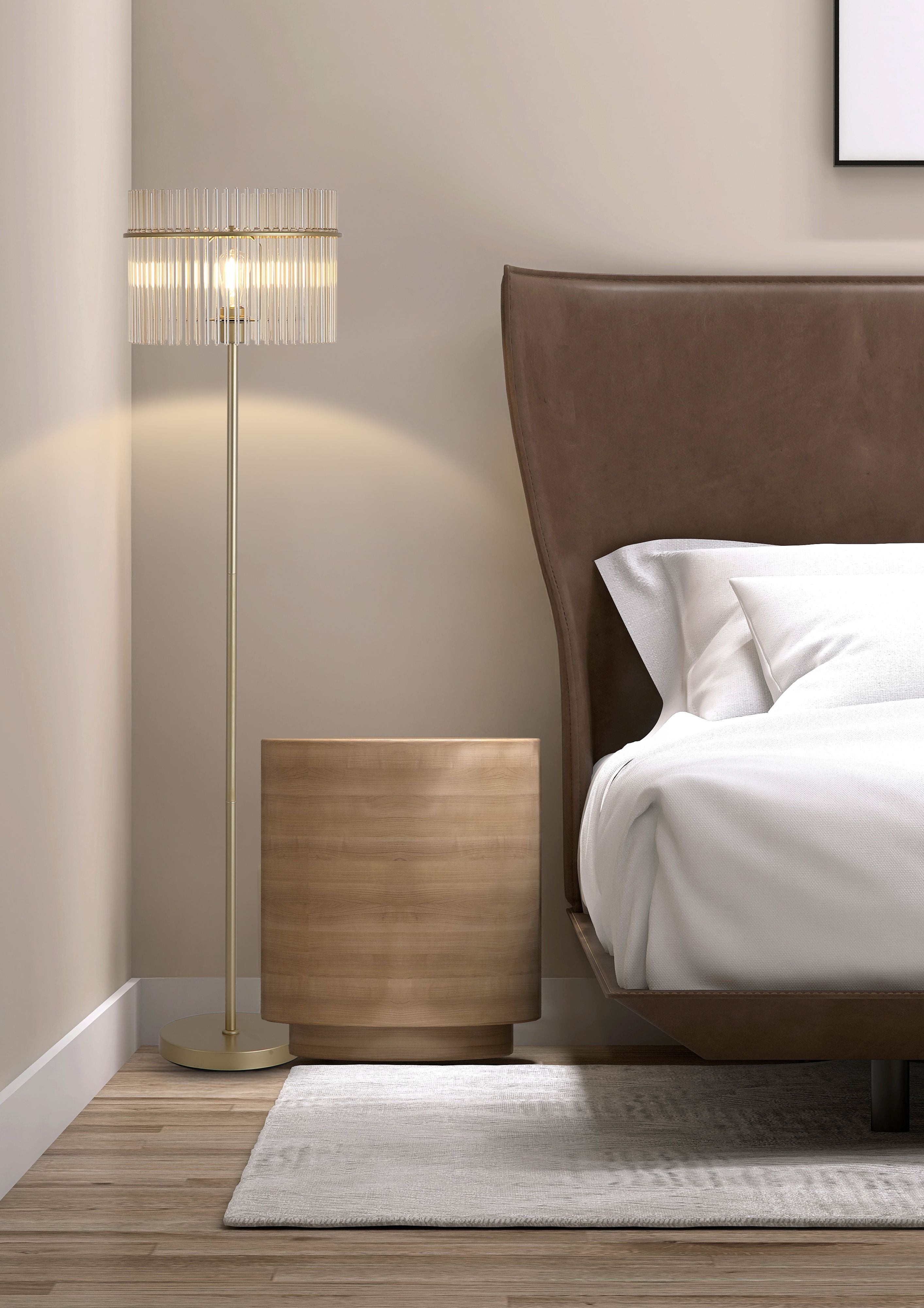Swizzle floor lamp in gold styled beside a bed in a bedroom, switched on next to a round wooden bedside table