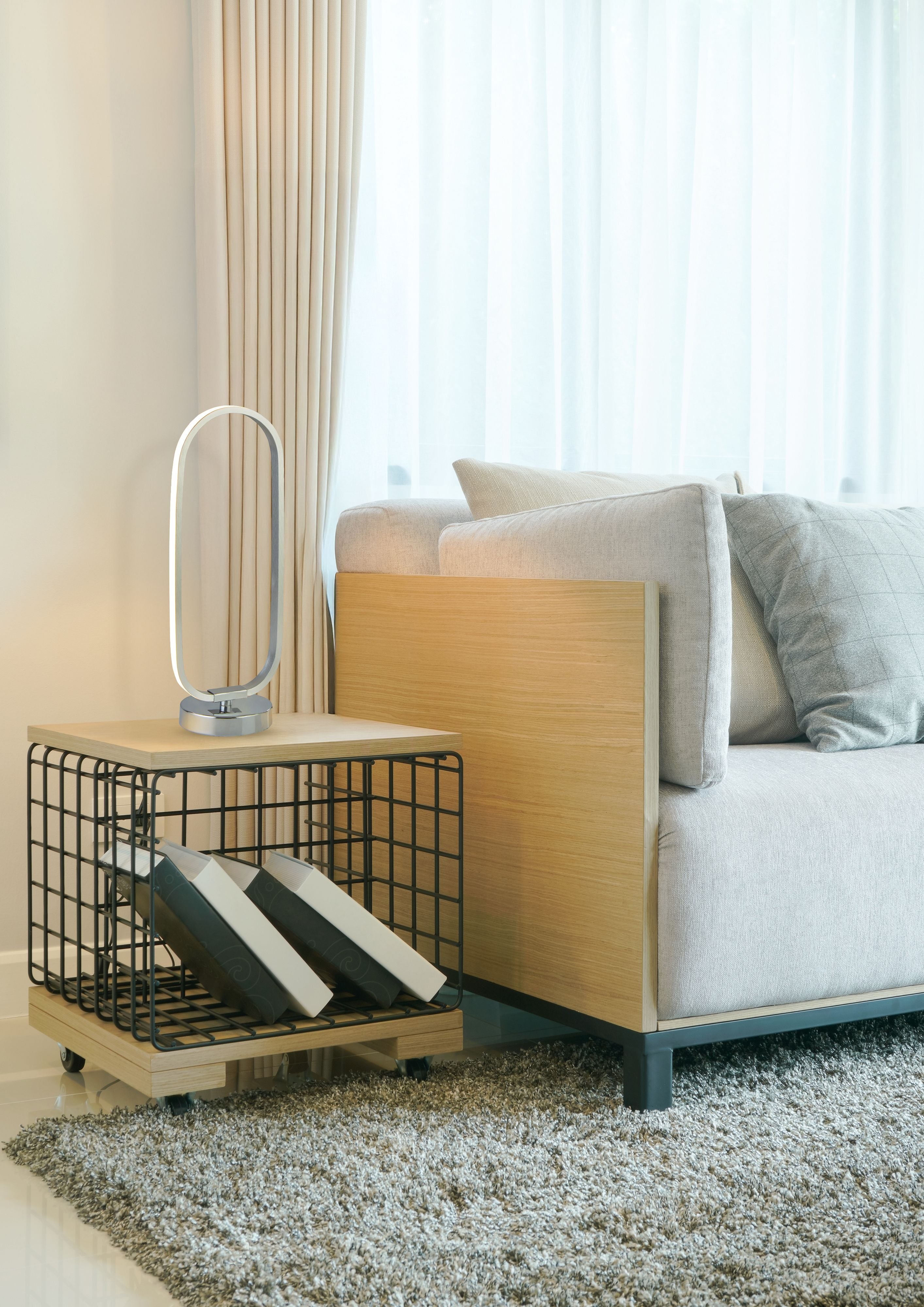 Liner chrome LED ring table lamp styled on a side table next to a wire basket of books and a sofa