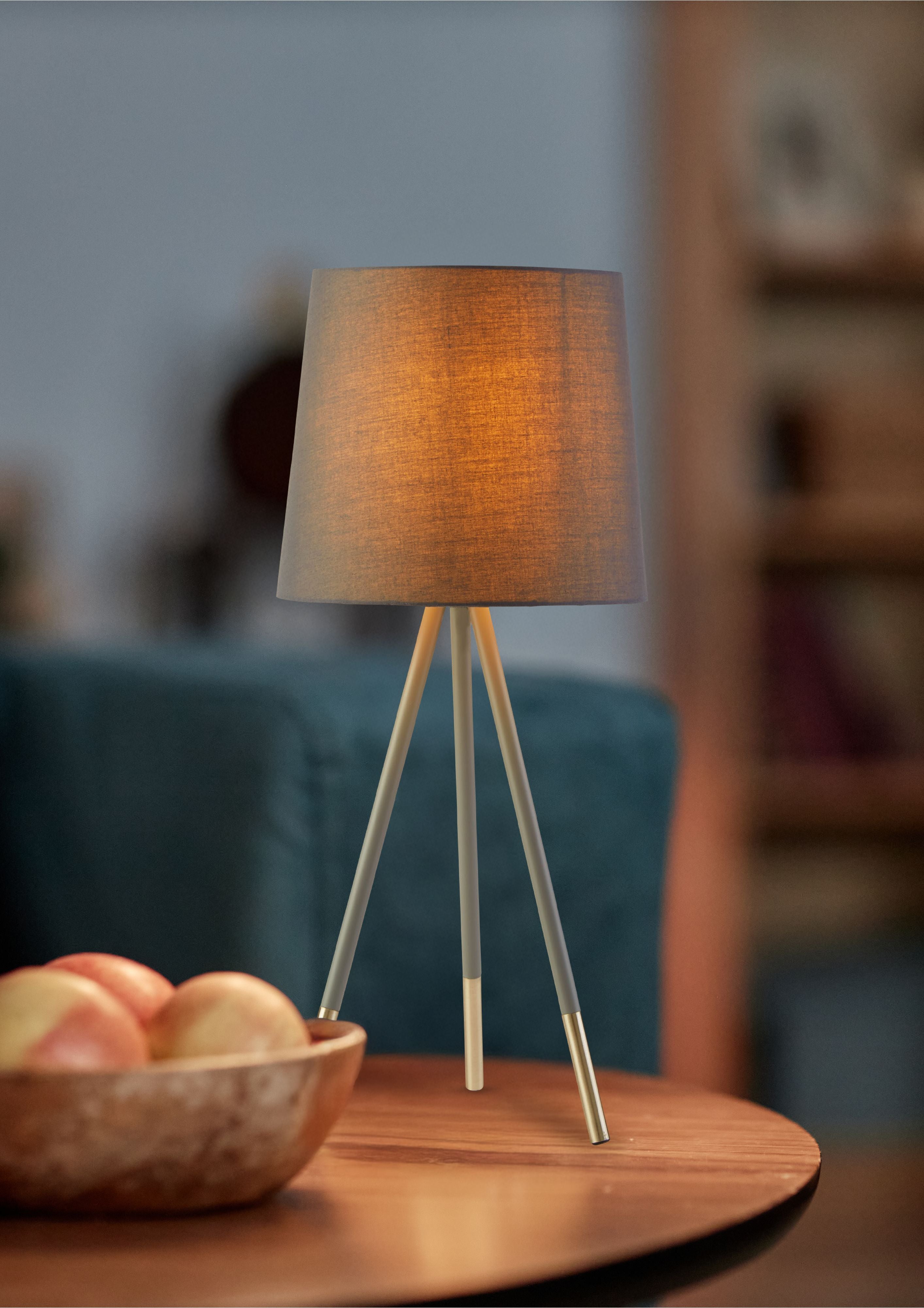 Rio table lamp lit on a round wooden side table next to a bowl of fruit, styled in a living room setting