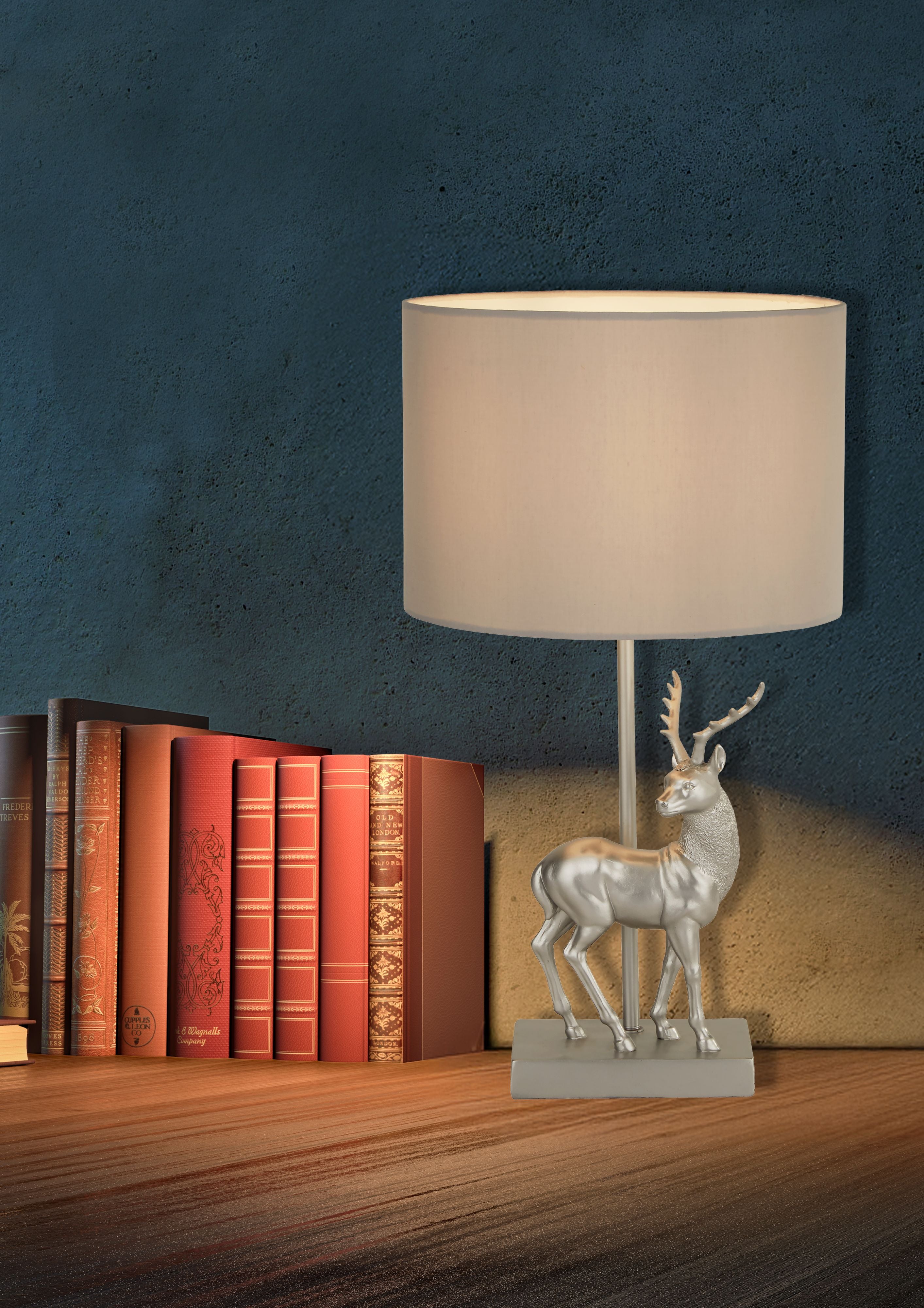 Searchlight Deer table lamp switched on with a warm glow, styled on a wooden shelf beside a row of red leather bound books against a dark teal mottled wall