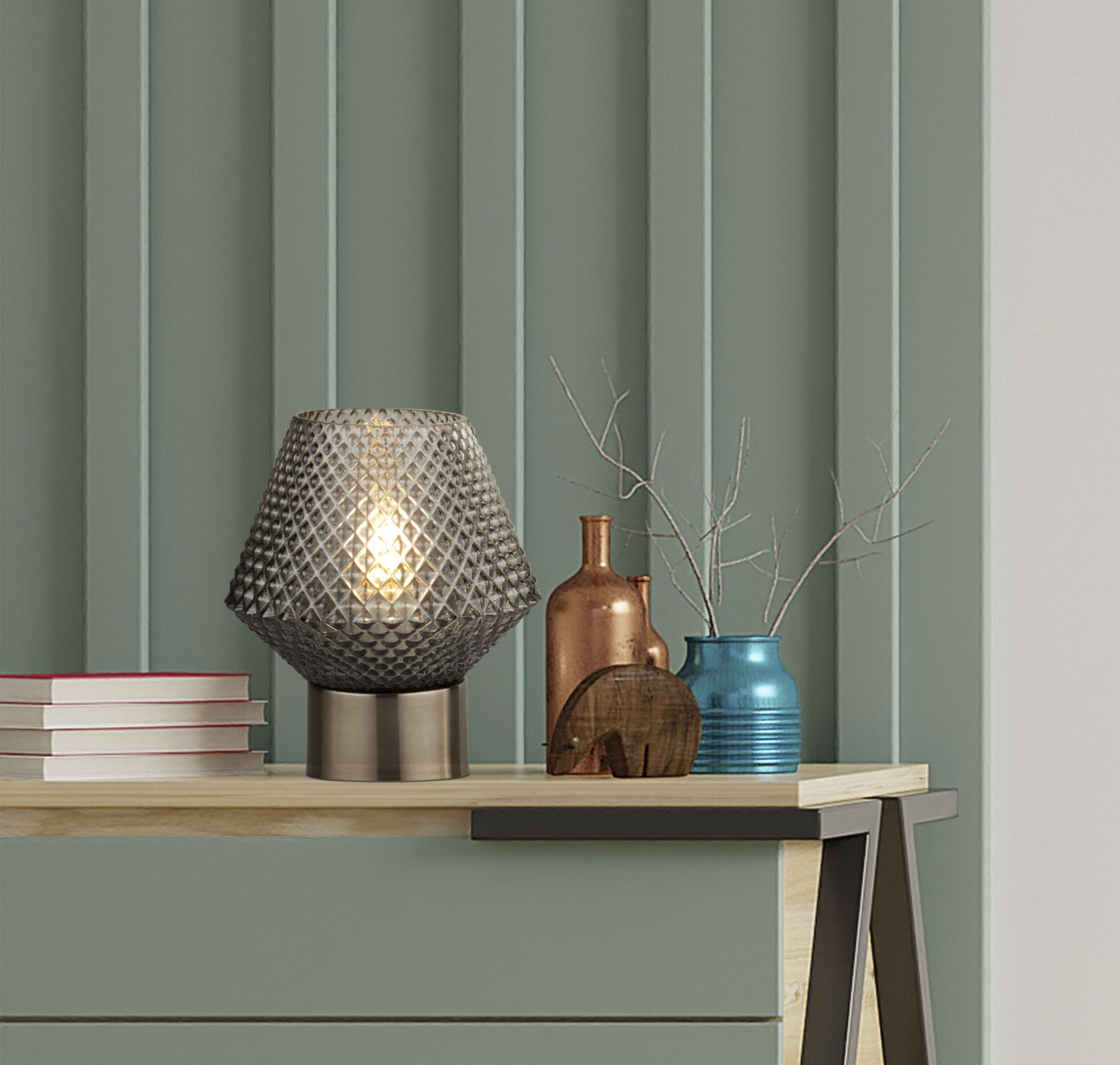 Searchlight Avia table lamp lit and styled on a wooden shelf beside books, small vases and a carved wooden ornament
