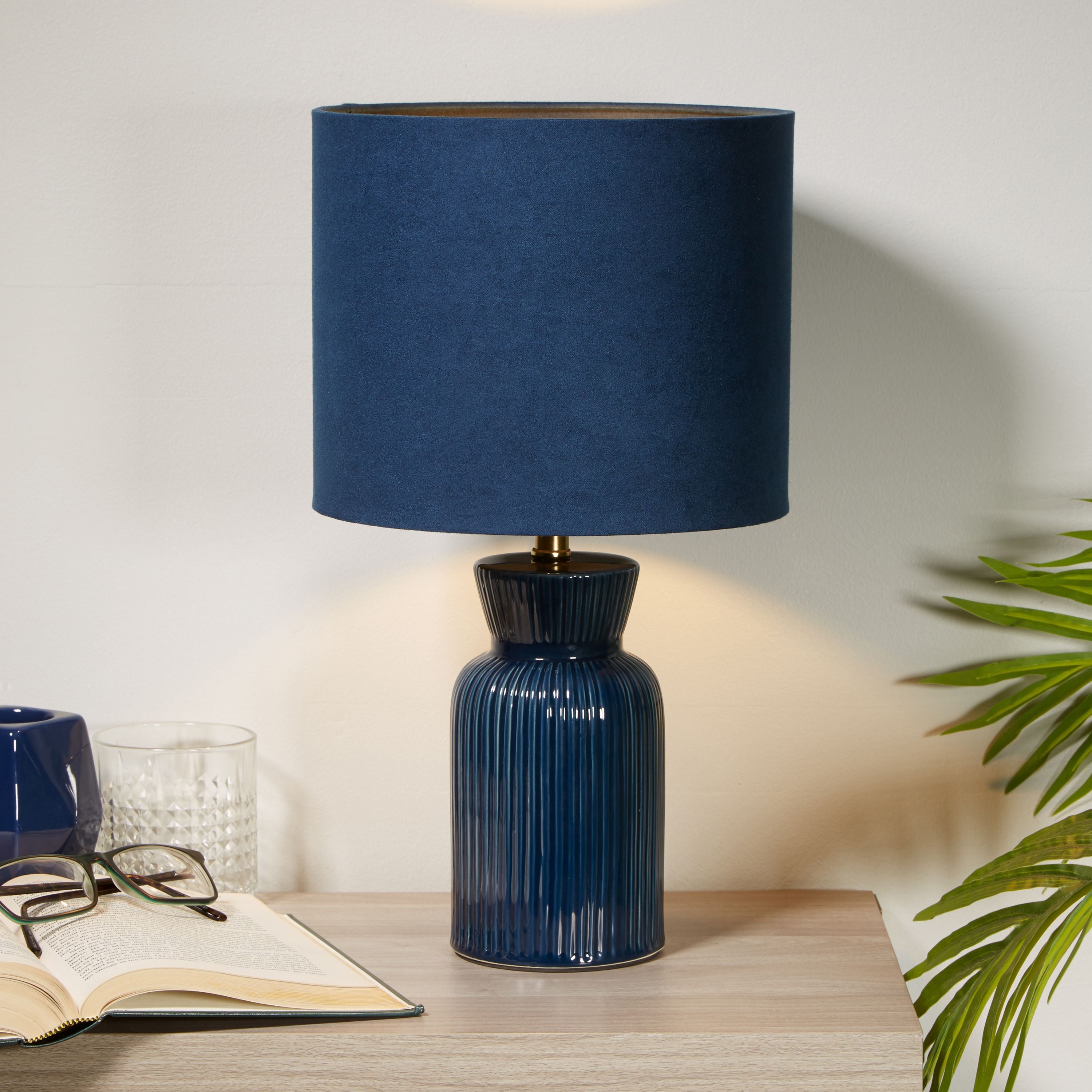 Navy ceramic table lamp styled on a light wood bedside table next to an open book, glasses, a glass tumbler and trailing green foliage