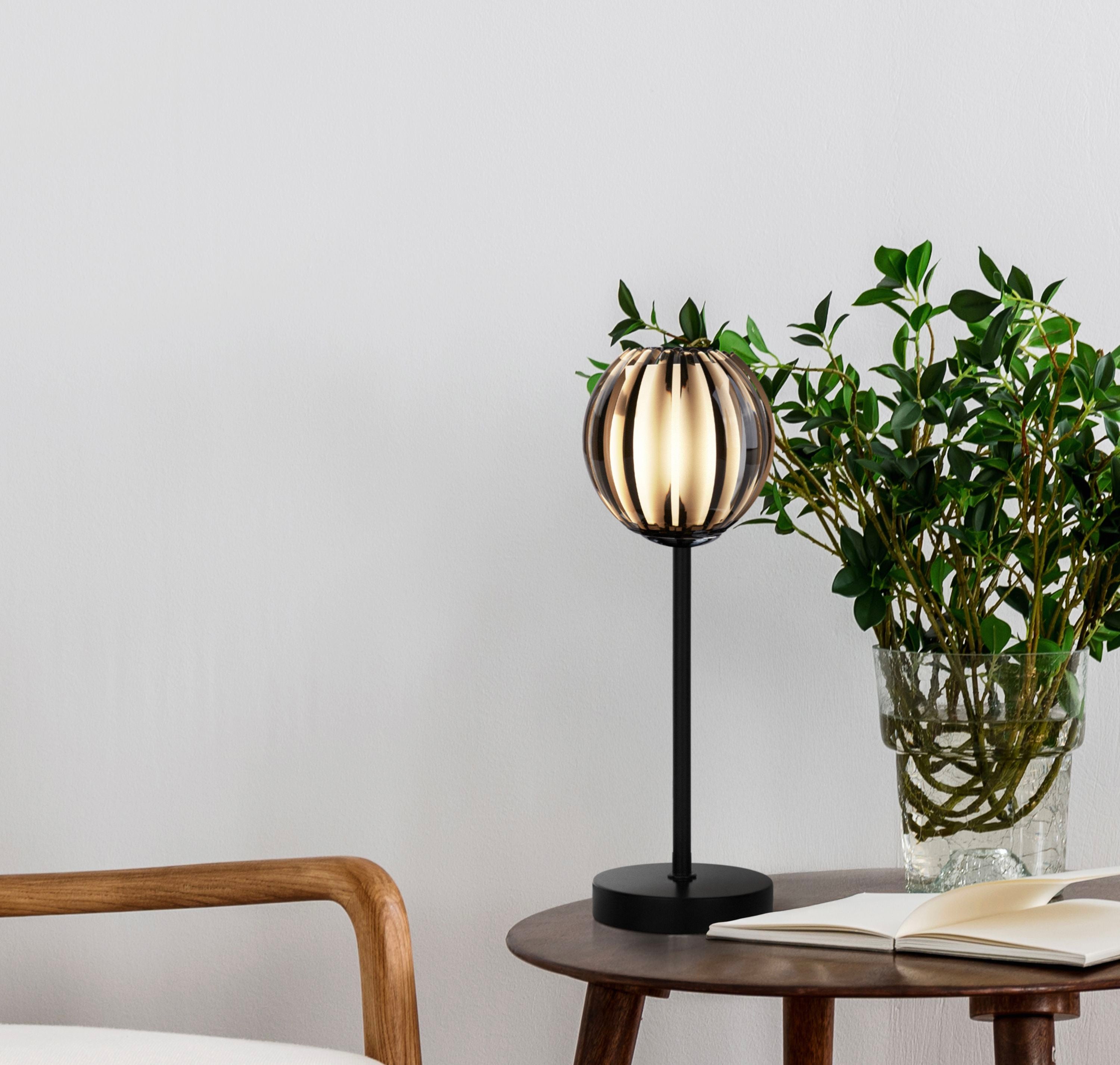 Blossom black table lamp with smoked acrylic shade styled on a side table next to a green plant and wooden armchair