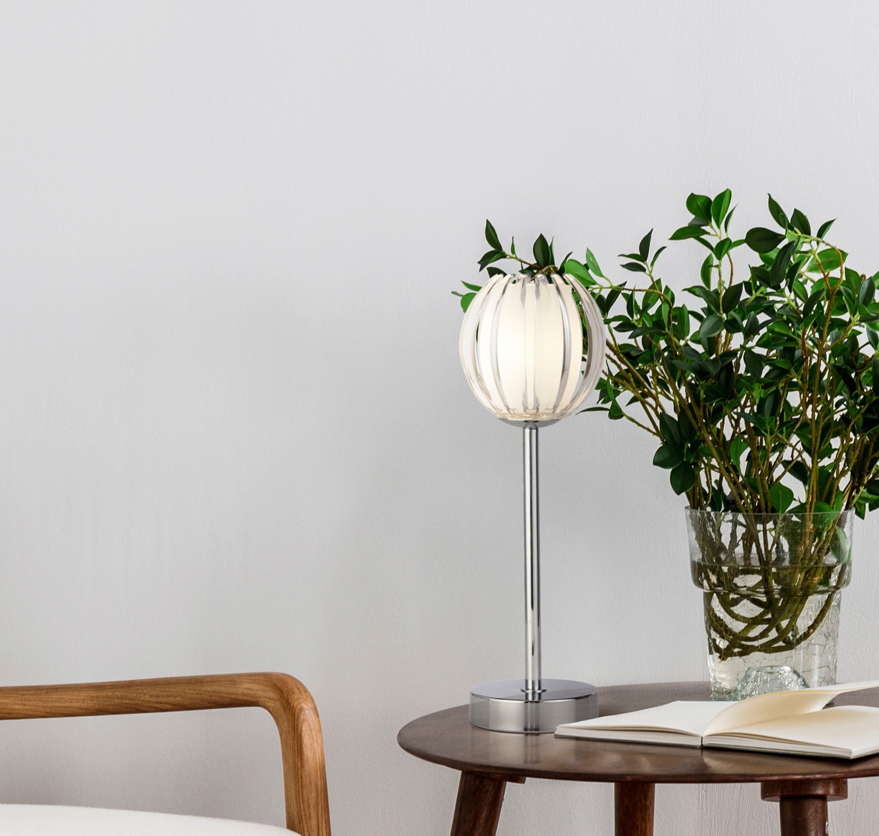 Searchlight Blossom table lamp with chrome stem and clear acrylic shade styled on a round wooden side table beside a green potted plant and an open book