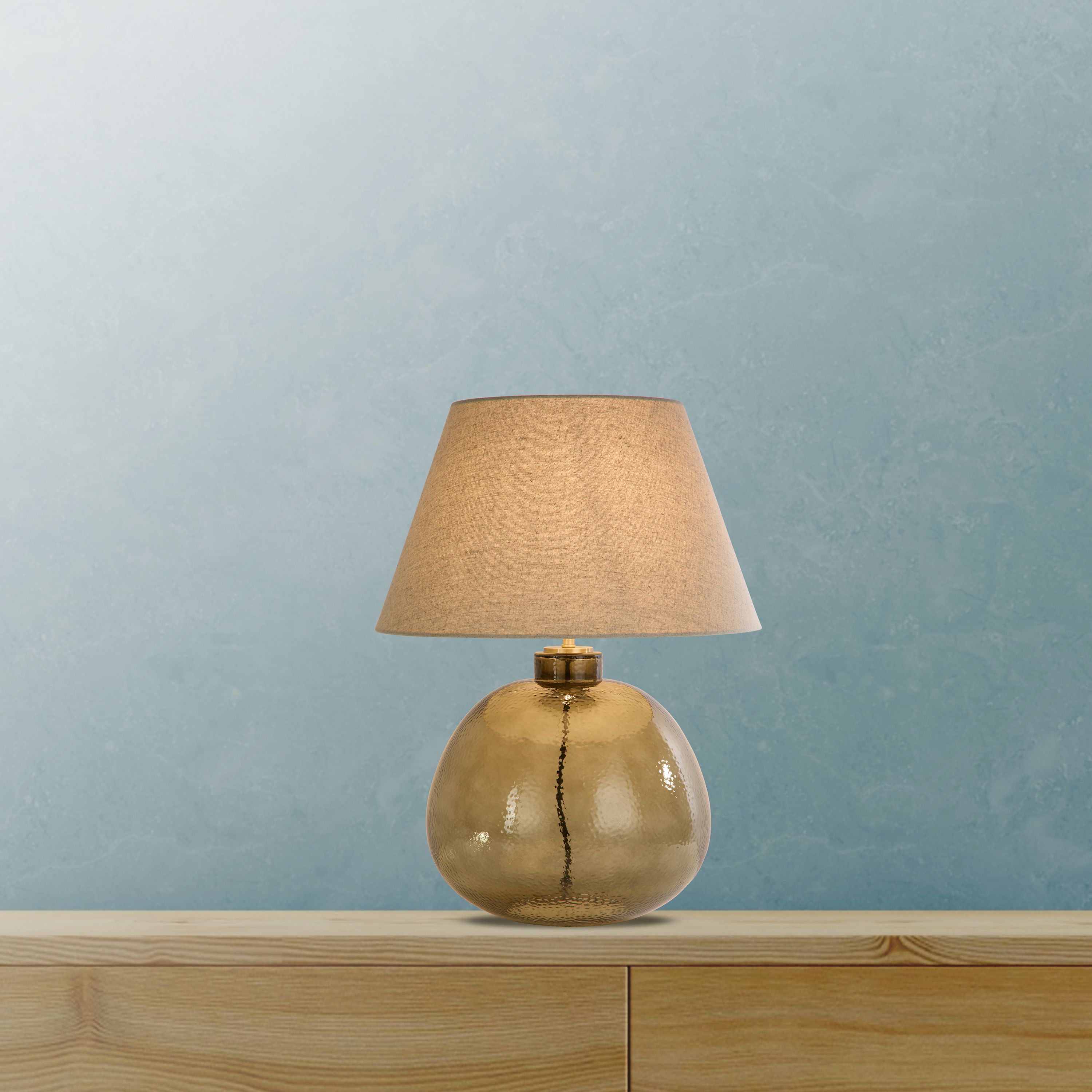 Lit table lamp with a round, ball shaped amber glass base and a beige tapered shade, styled against a blue mottled wall on a wooden shelf
