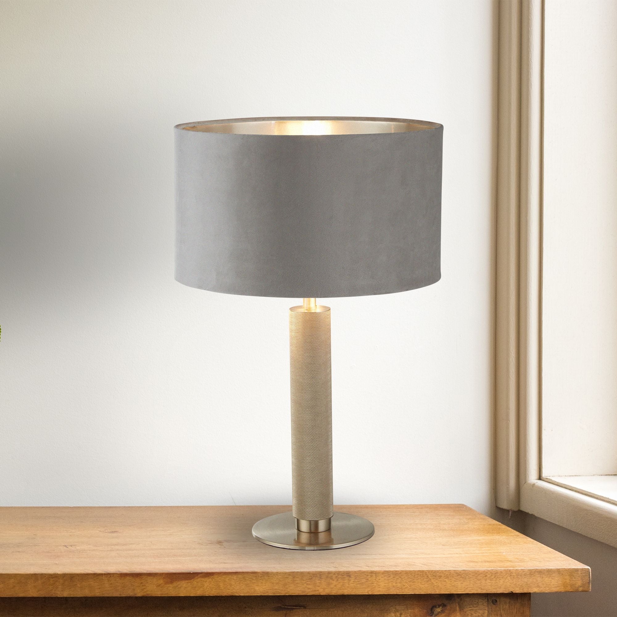 London table lamp with grey velvet shade and knurled metal column styled switched on, on a wooden sideboard beside a window