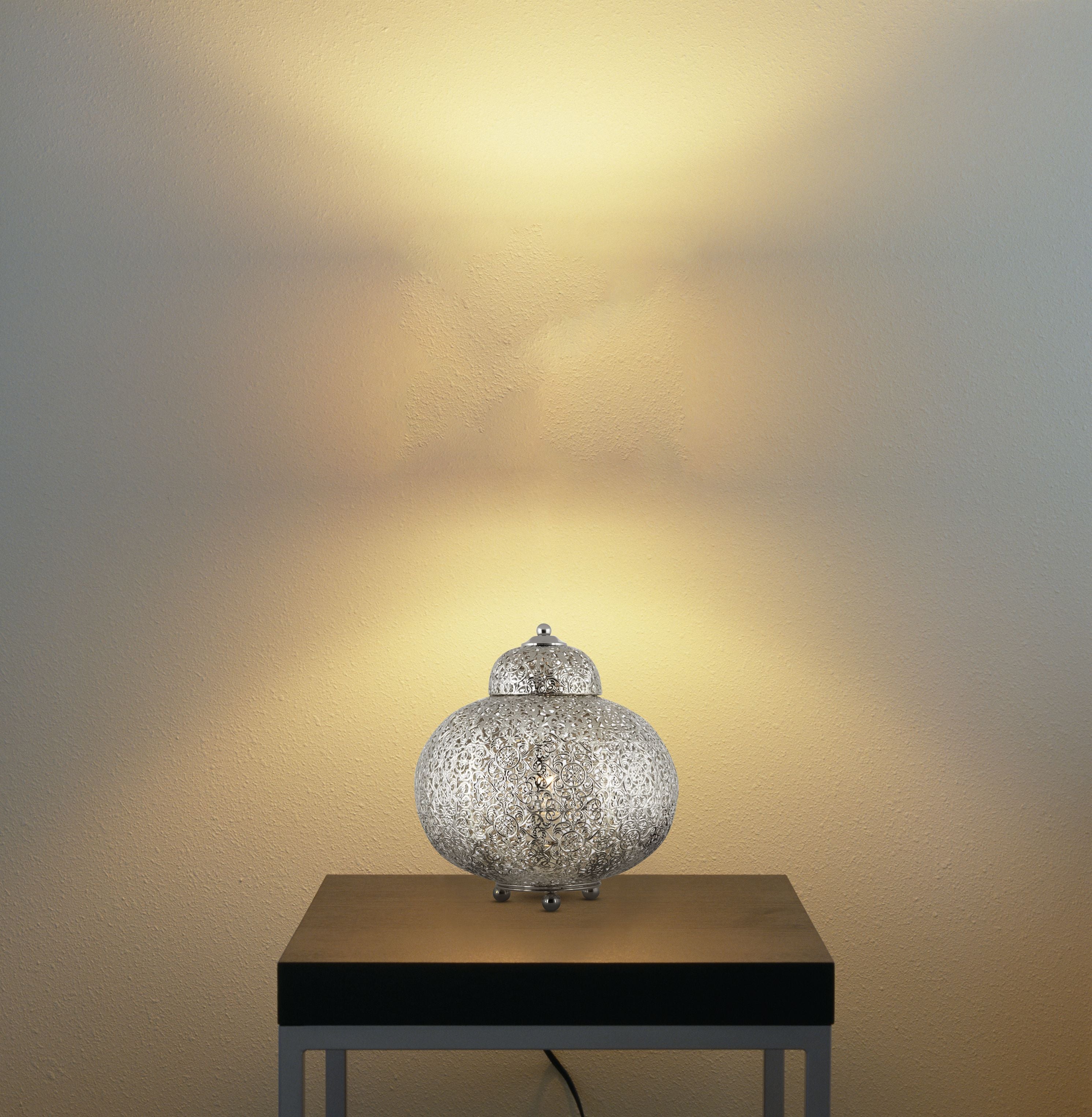 Fretwork table lamp in nickel styled on a bedside table, lit against a wall with a warm glow