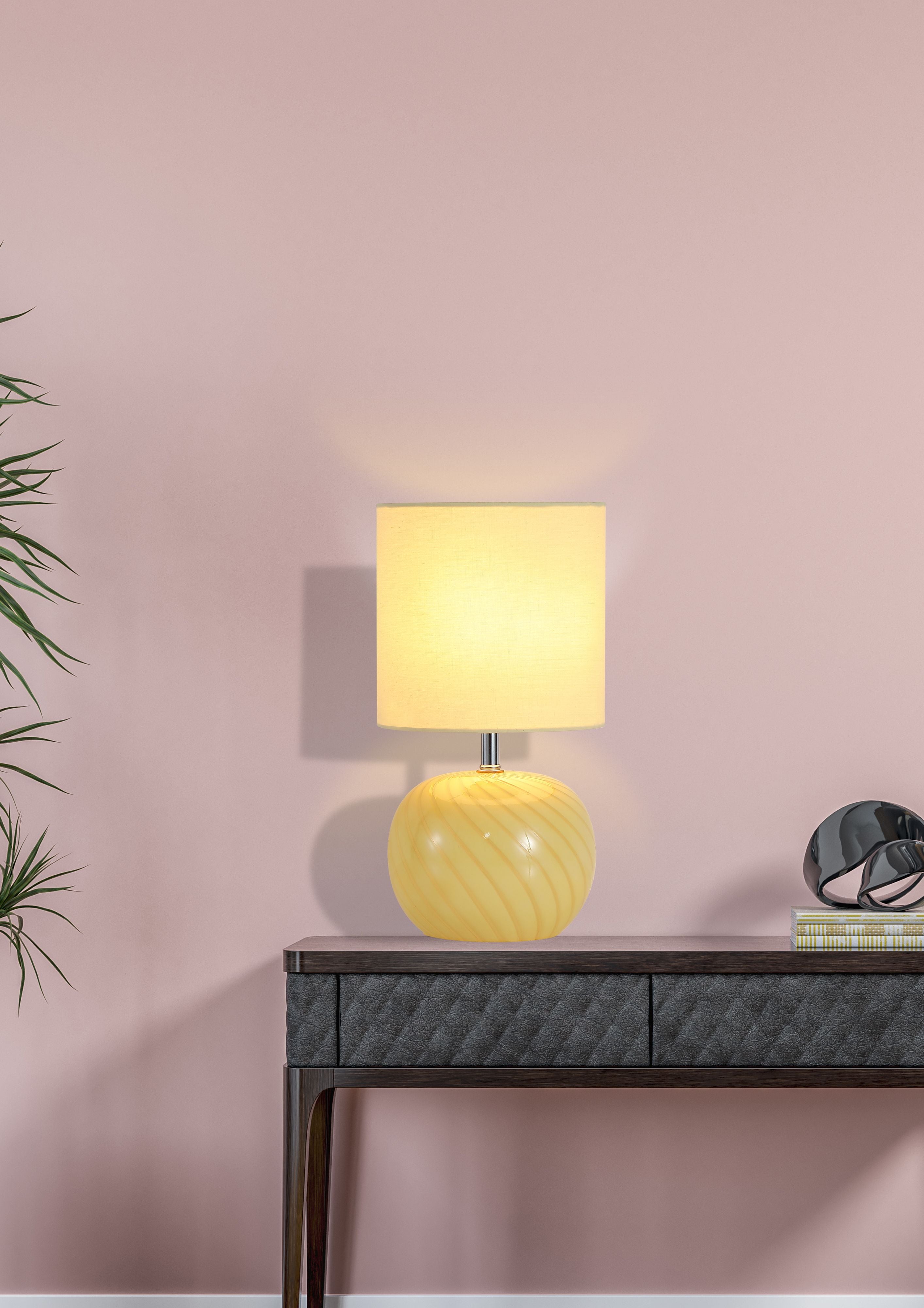 Fete yellow glass table lamp styled on a dark console table against a pink wall, lit with a warm glow, with a plant and decorative sphere nearby