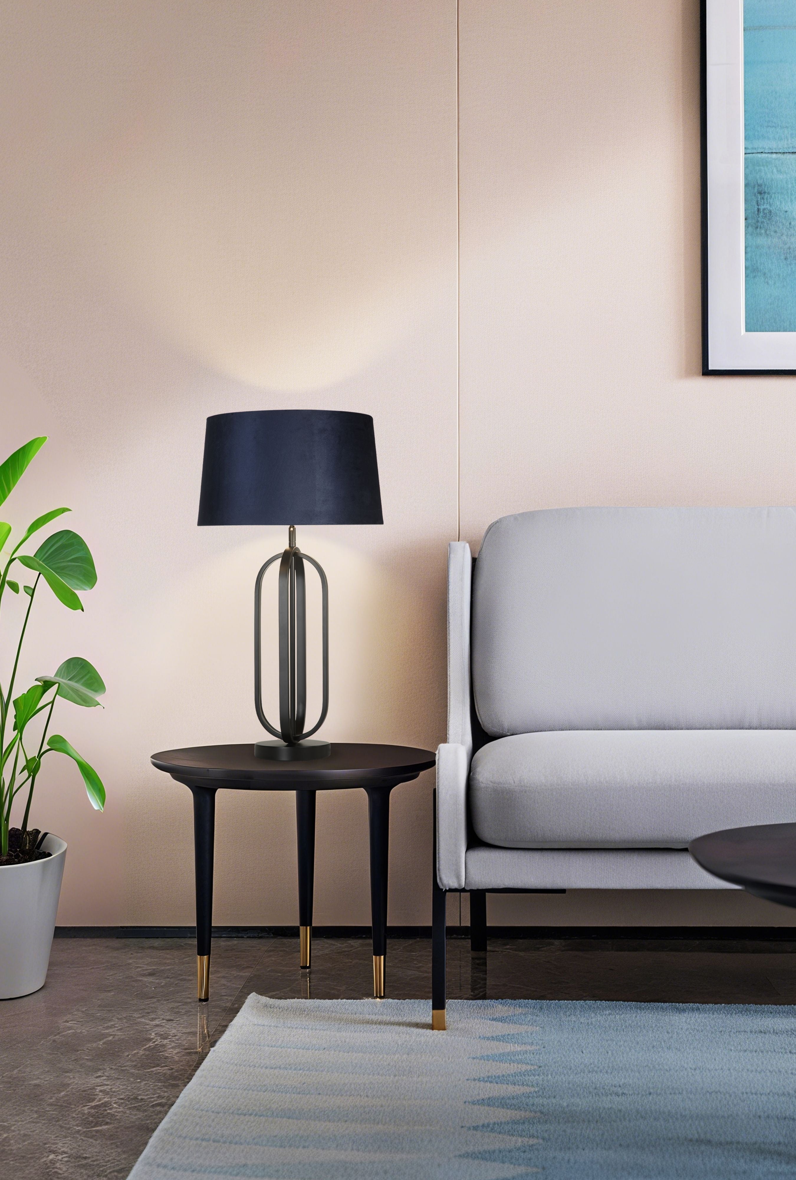 The lamp styled on a round side table beside a light grey armchair and a potted plant, its navy velvet shade glowing warm light in a blush pink panelled room