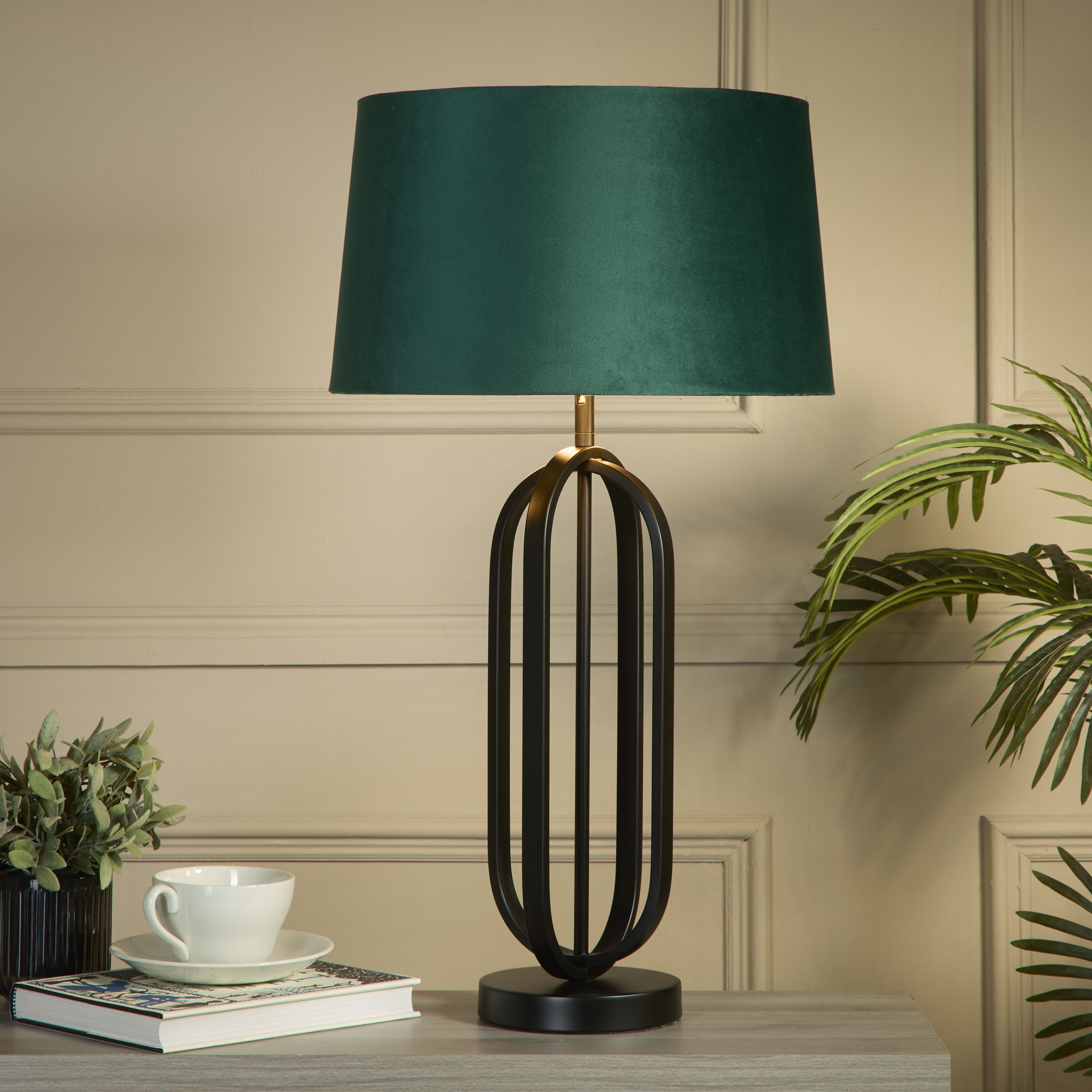 The lamp styled on a dark wood side table against a beige panelled wall, with a teacup and saucer, a book and potted plants either side, its emerald velvet shade glowing warm light