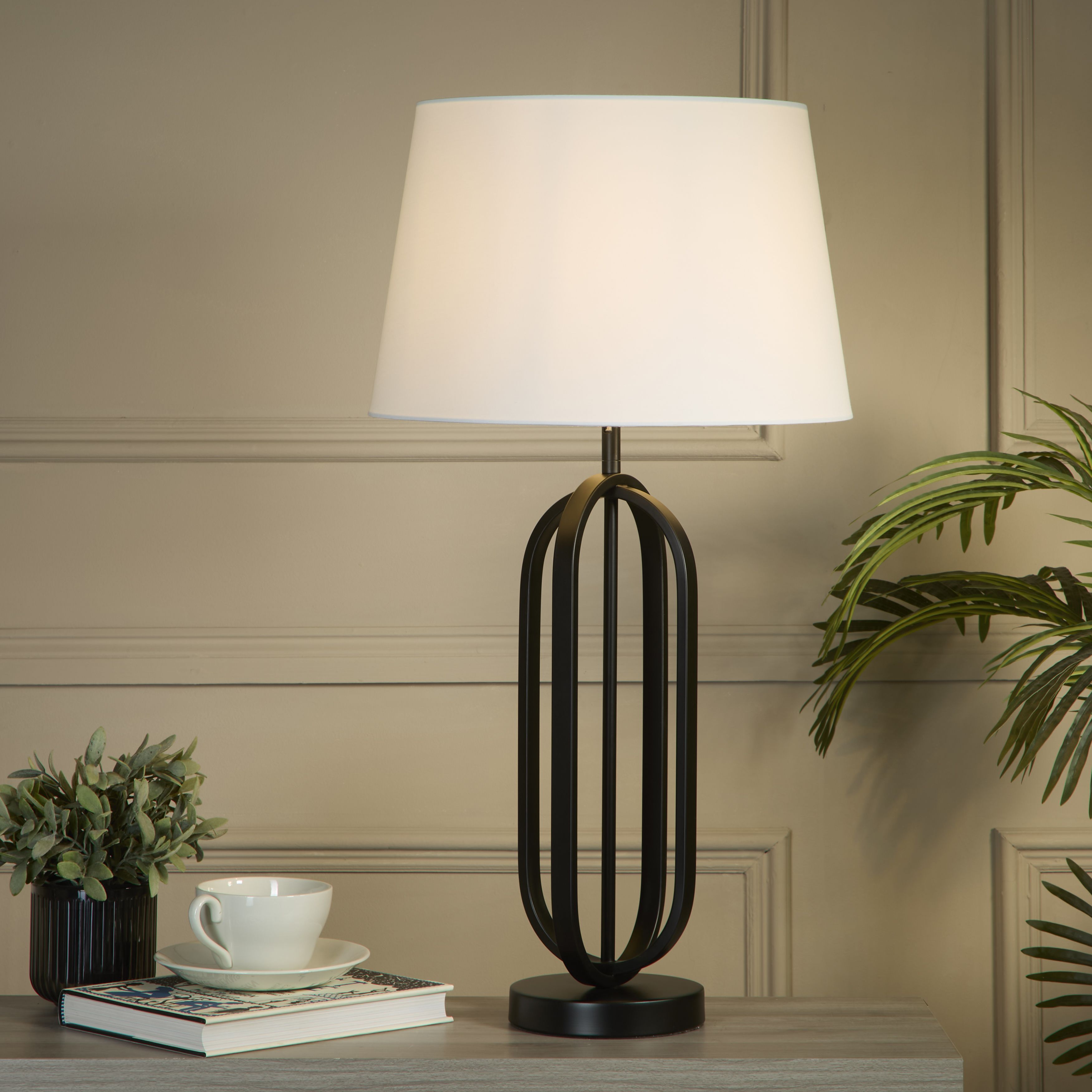 The lamp styled on a grey table against a taupe panelled wall, with a teacup and saucer, a book and potted plants either side, its white shade glowing warm light