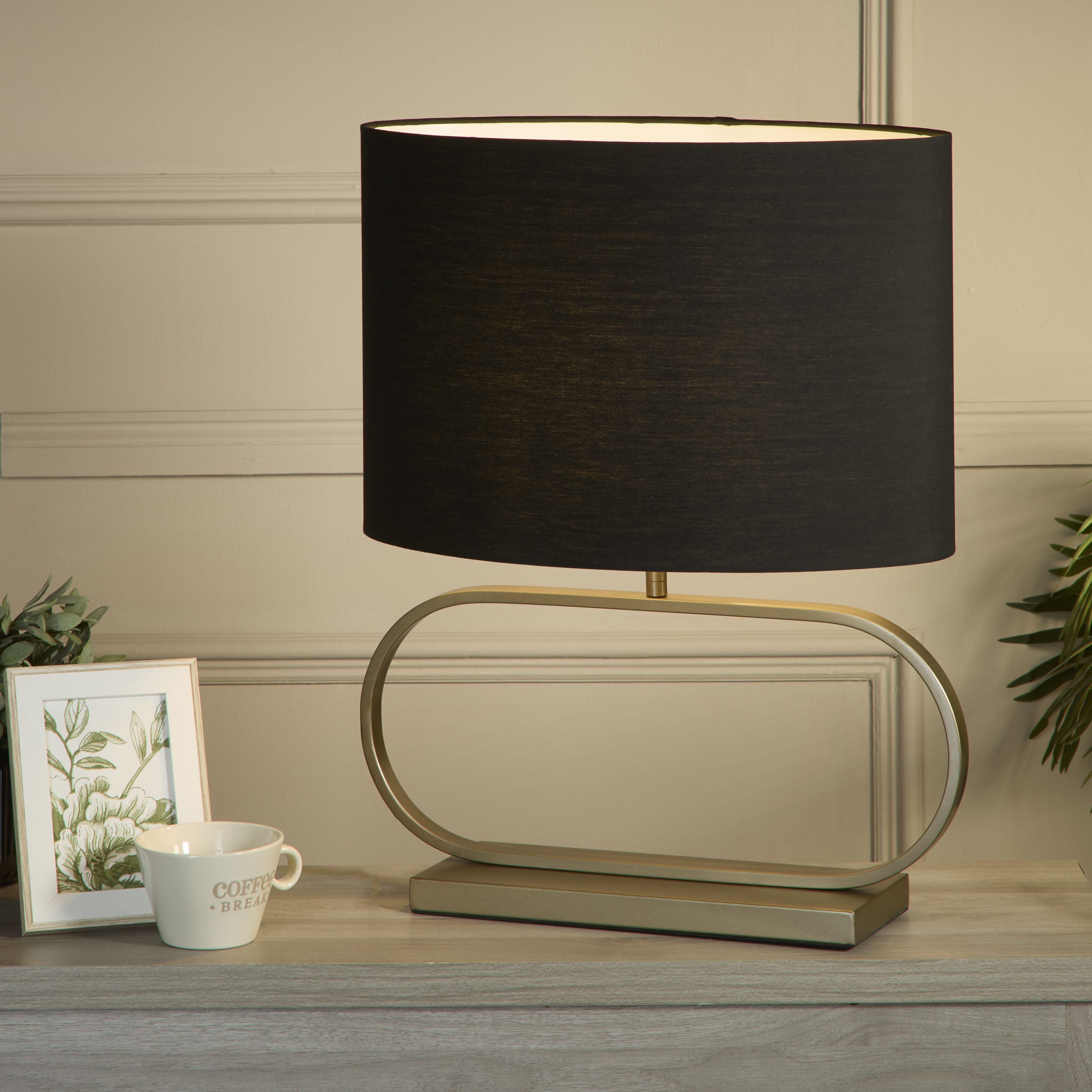 Sleek table lamp in bronze with a black shade styled on a living room sideboard beside a picture frame, a cup, and a small vase of foliage, shade glowing warm white.