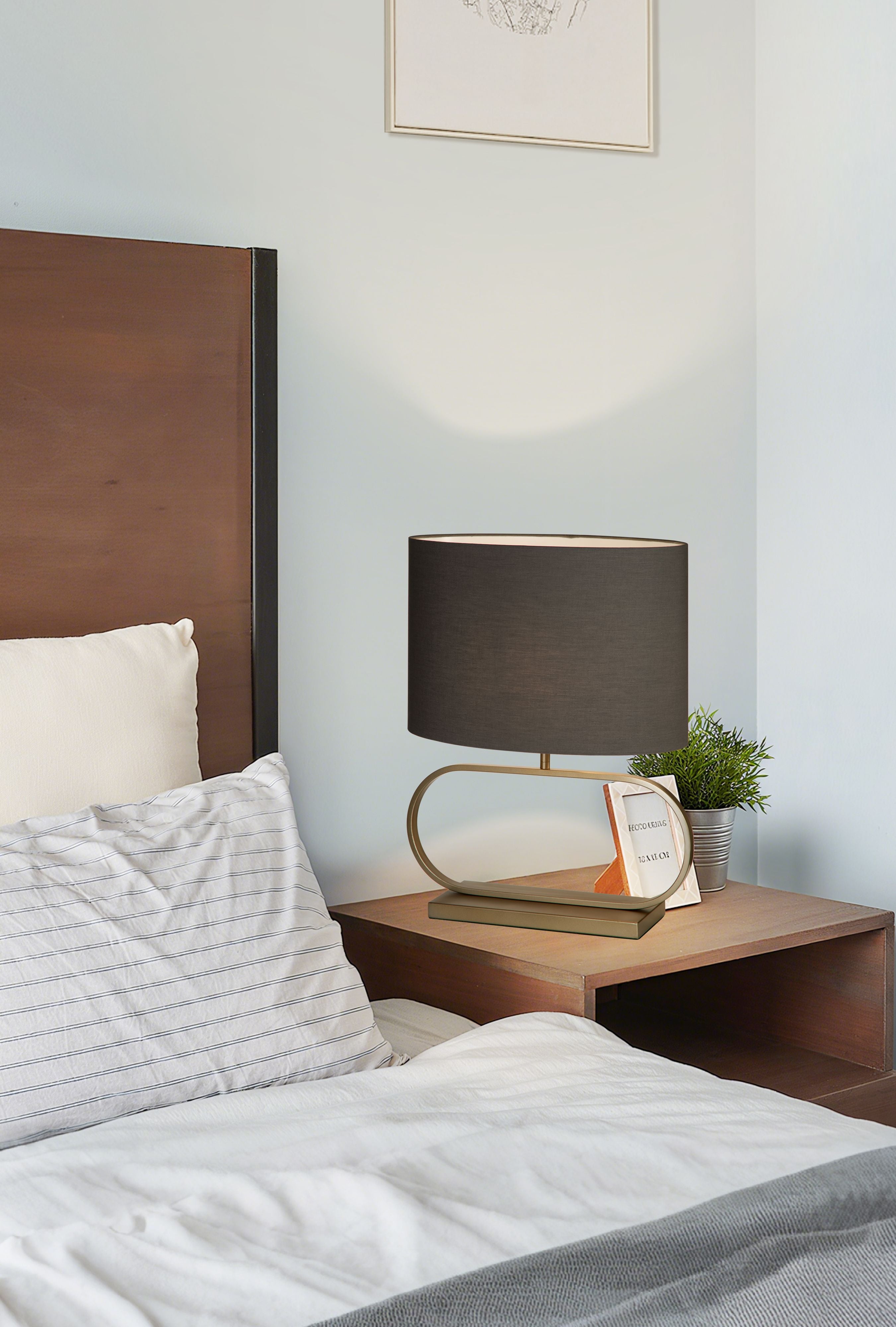 Sleek table lamp in bronze with a grey shade styled on a bedroom nightstand beside a picture frame and a potted plant, shade glowing warm white.