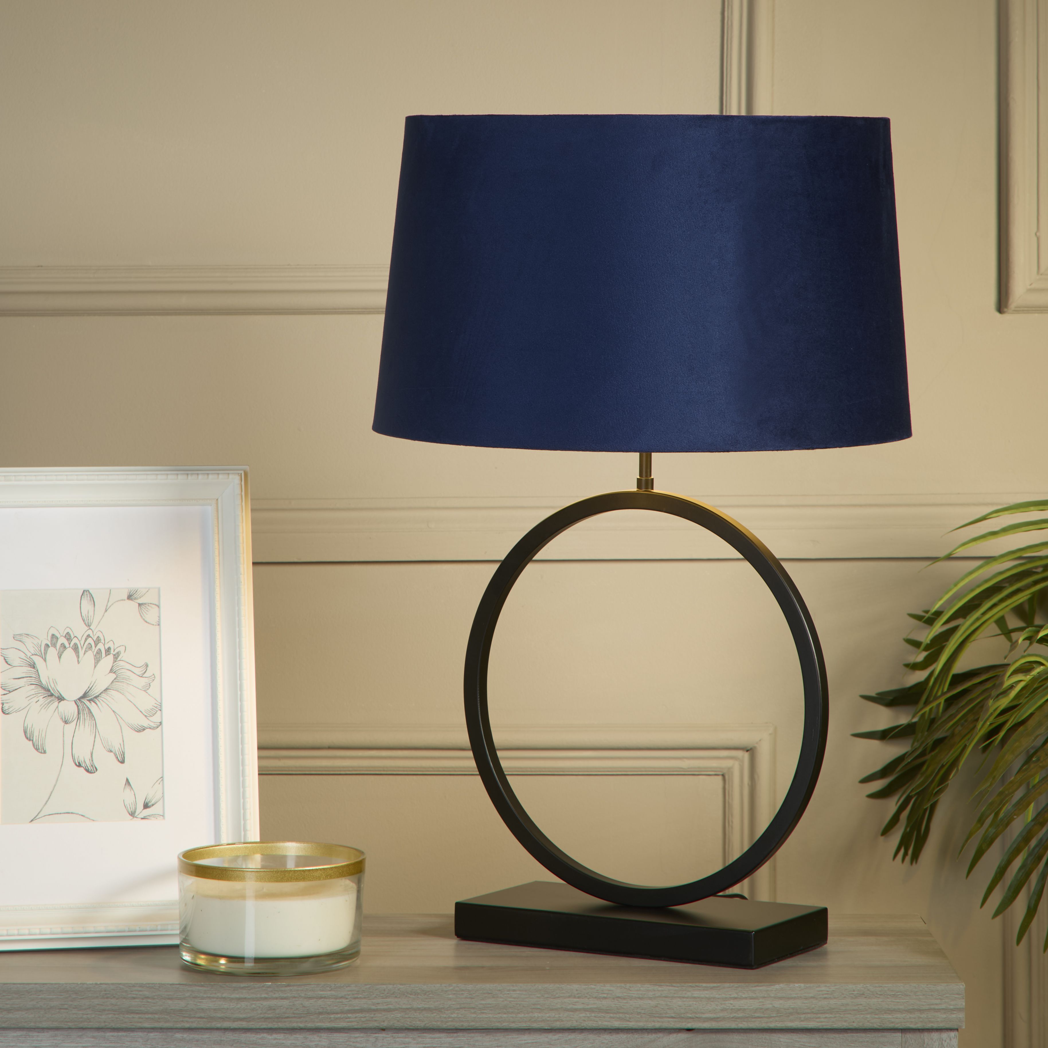 Elegant Table Lamp with navy velvet shade styled on a wooden bedside table next to a framed print, candle and a leafy plant