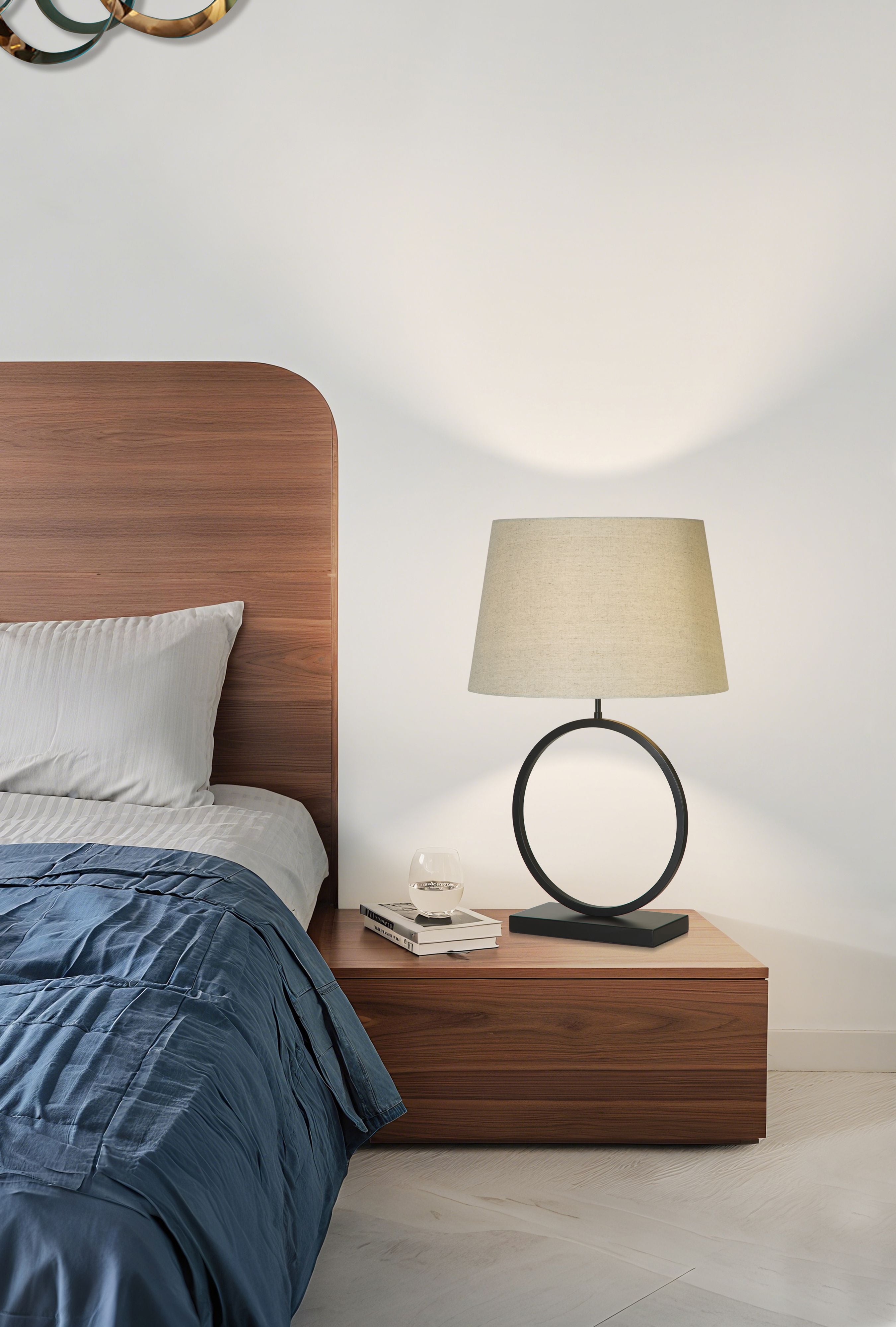 Elegant Table Lamp with natural linen shade lit up on a wooden bedside table next to a water glass and stacked books, walnut headboard and blue bedding in the background
