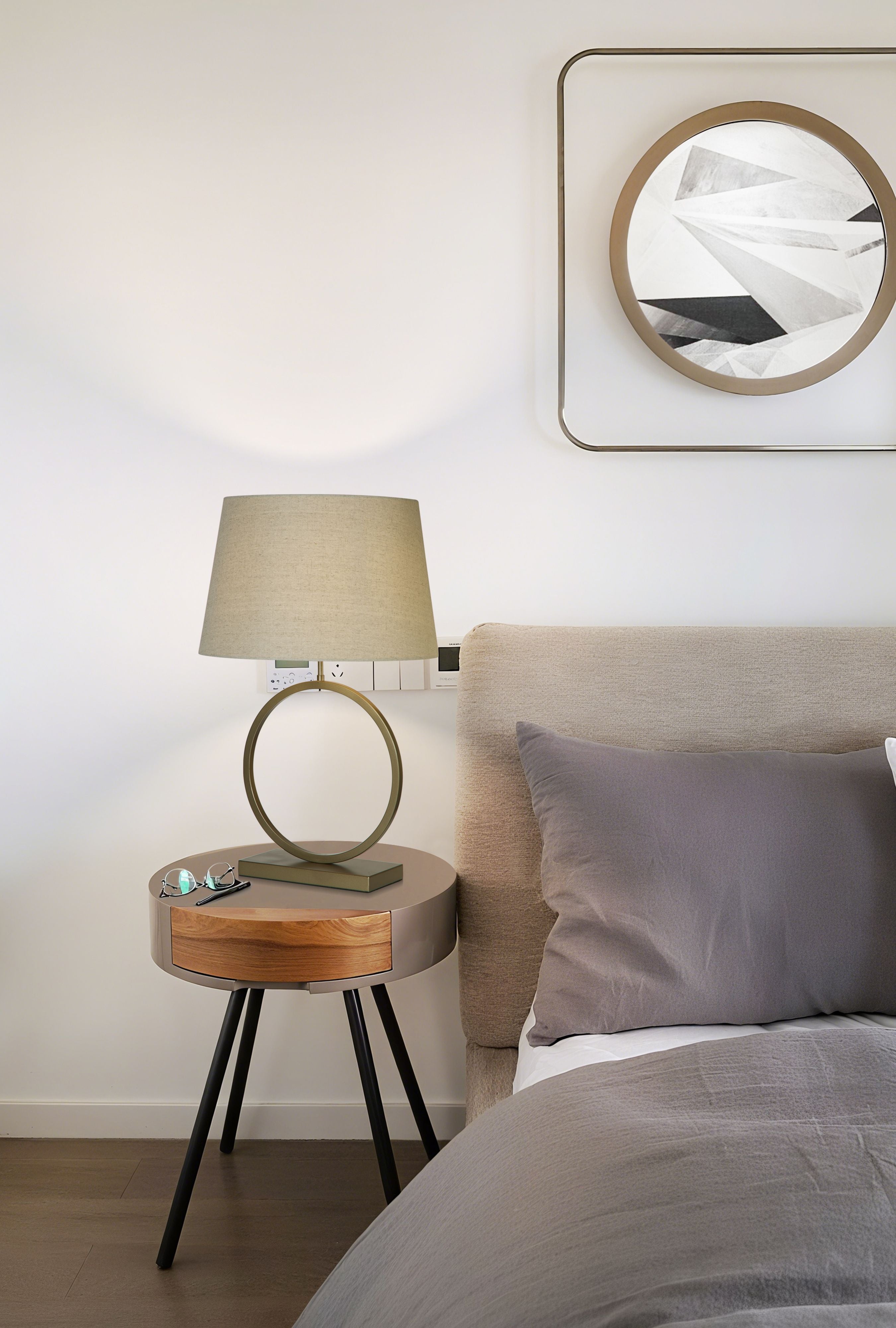 Elegant table lamp with natural linen shade, switched on, styled on a round bedside table next to a bed with a framed print above