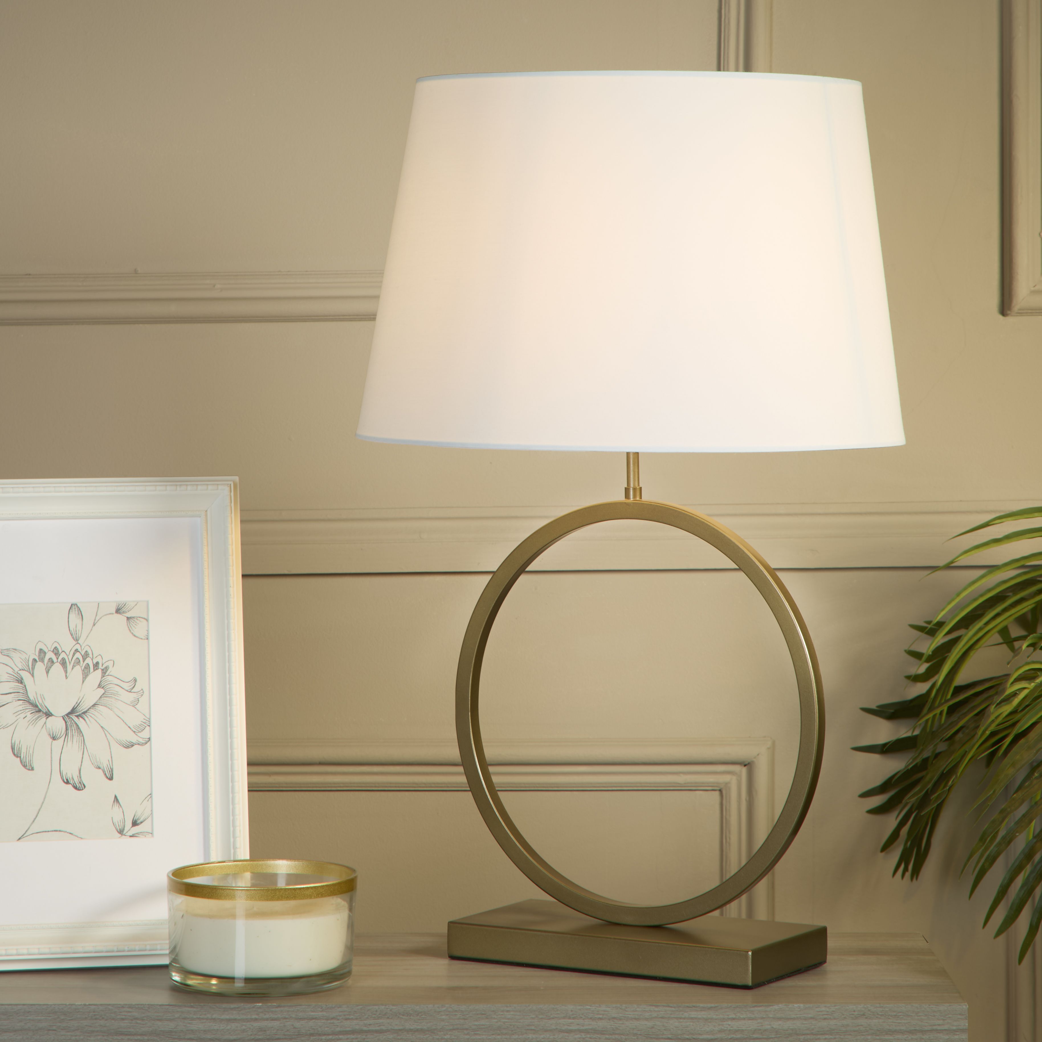 Elegant table lamp with white cotton shade switched on, styled on a console table with a framed botanical print, candle and trailing plant