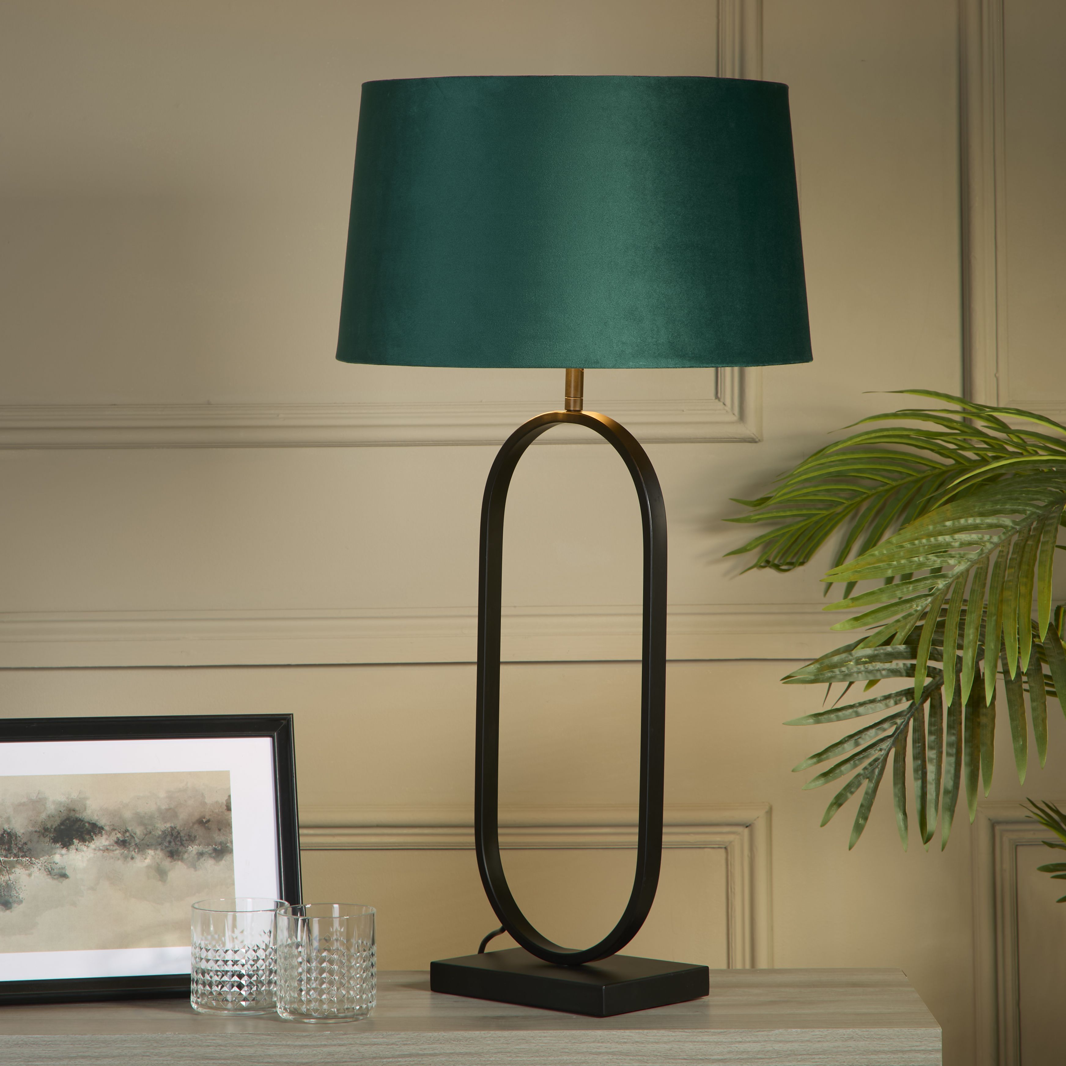 Modish table lamp with an emerald velvet shade styled on a light console table beside a framed print, two cut glass tumblers and a potted palm, shown switched on