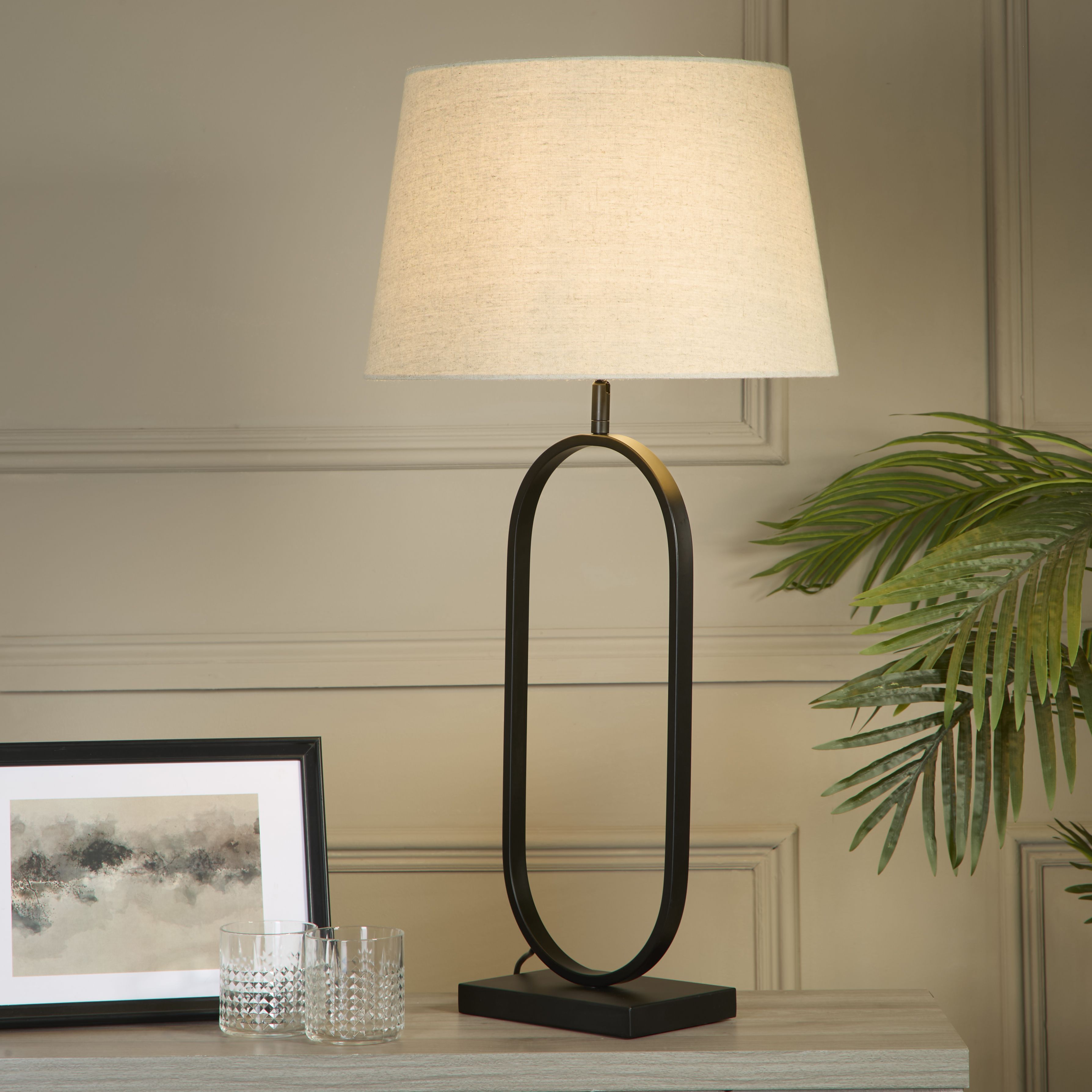 Modish table lamp with a natural linen shade styled on a light console table beside a framed print, two cut glass tumblers and a potted palm, shown switched on