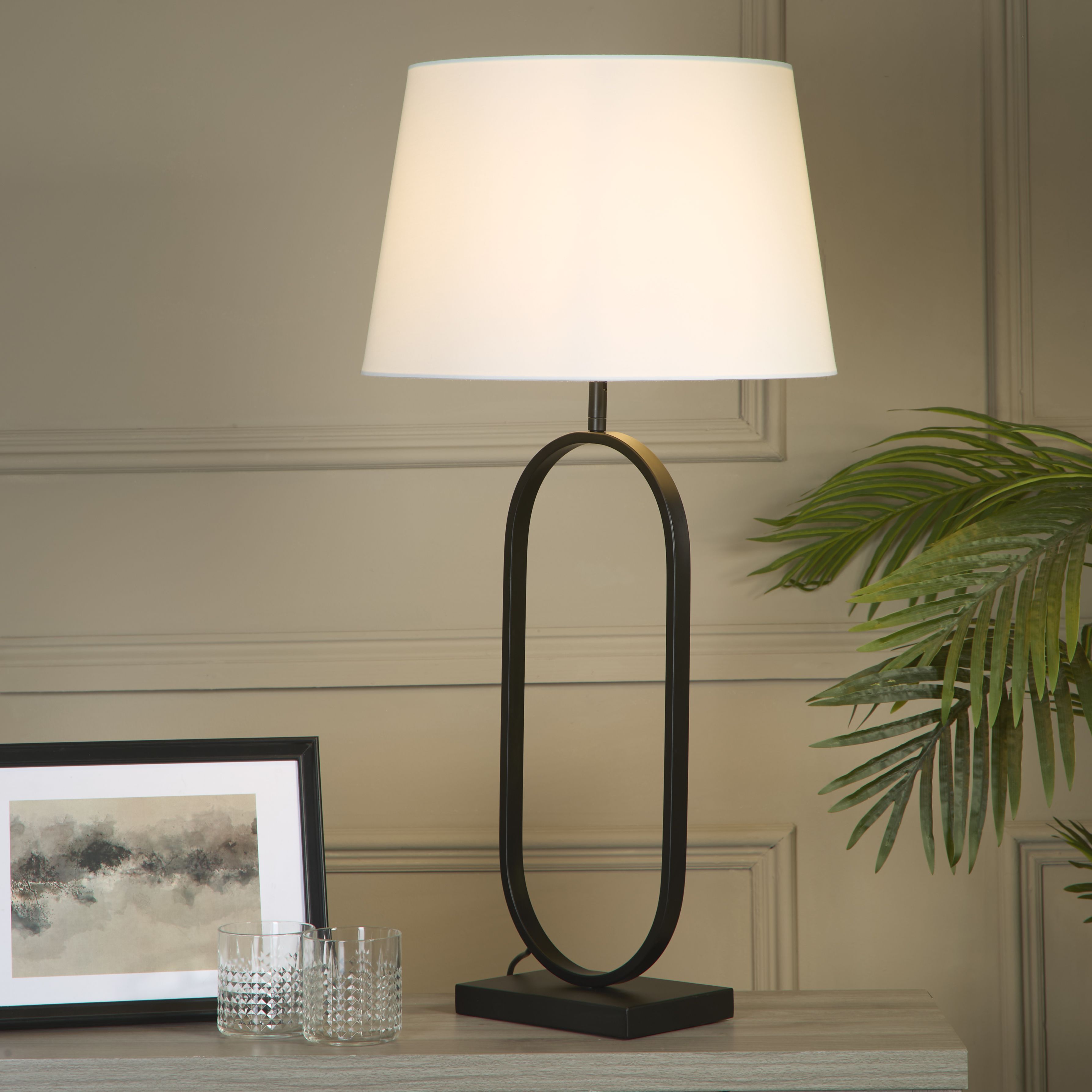 Modish table lamp with a white cotton shade styled on a light console table beside a framed print, two cut glass tumblers and a potted palm, shown switched on