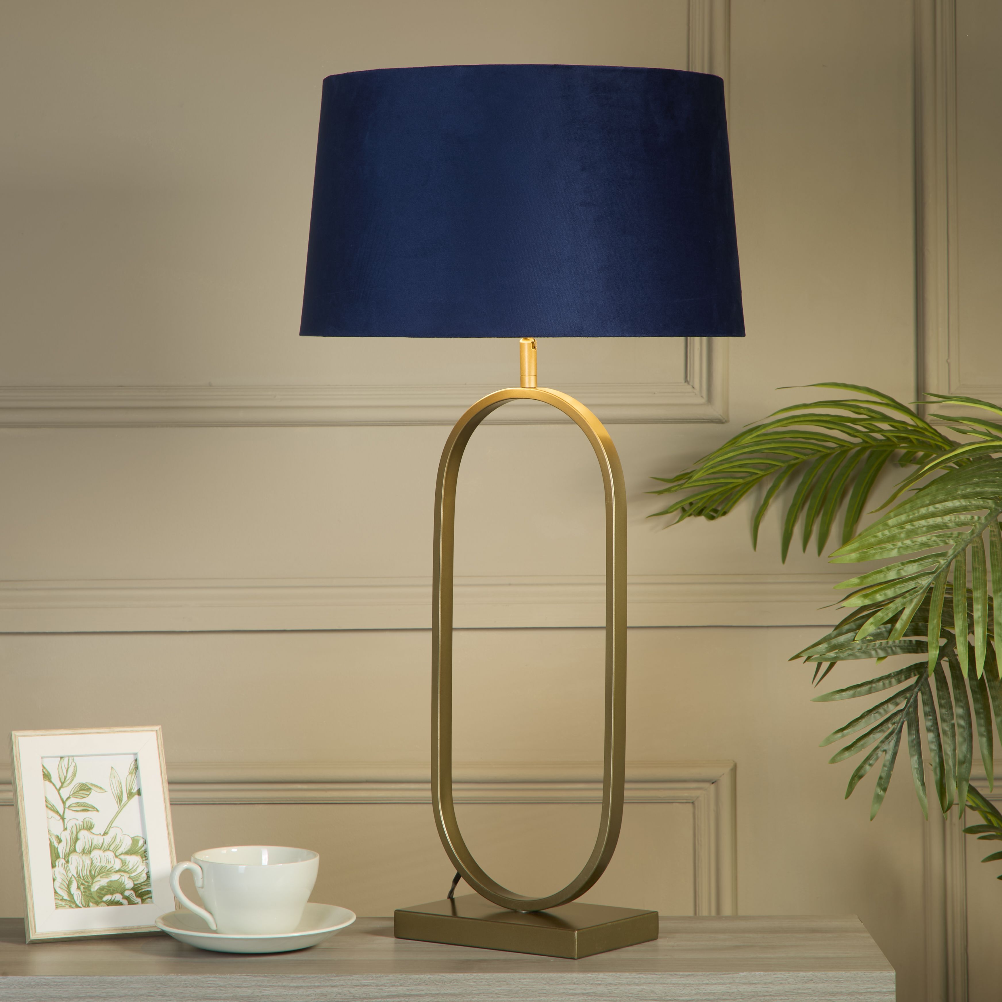 Modish table lamp with a navy velvet shade styled on a light console table beside a framed print, a cup and saucer, and a potted palm, shown switched on