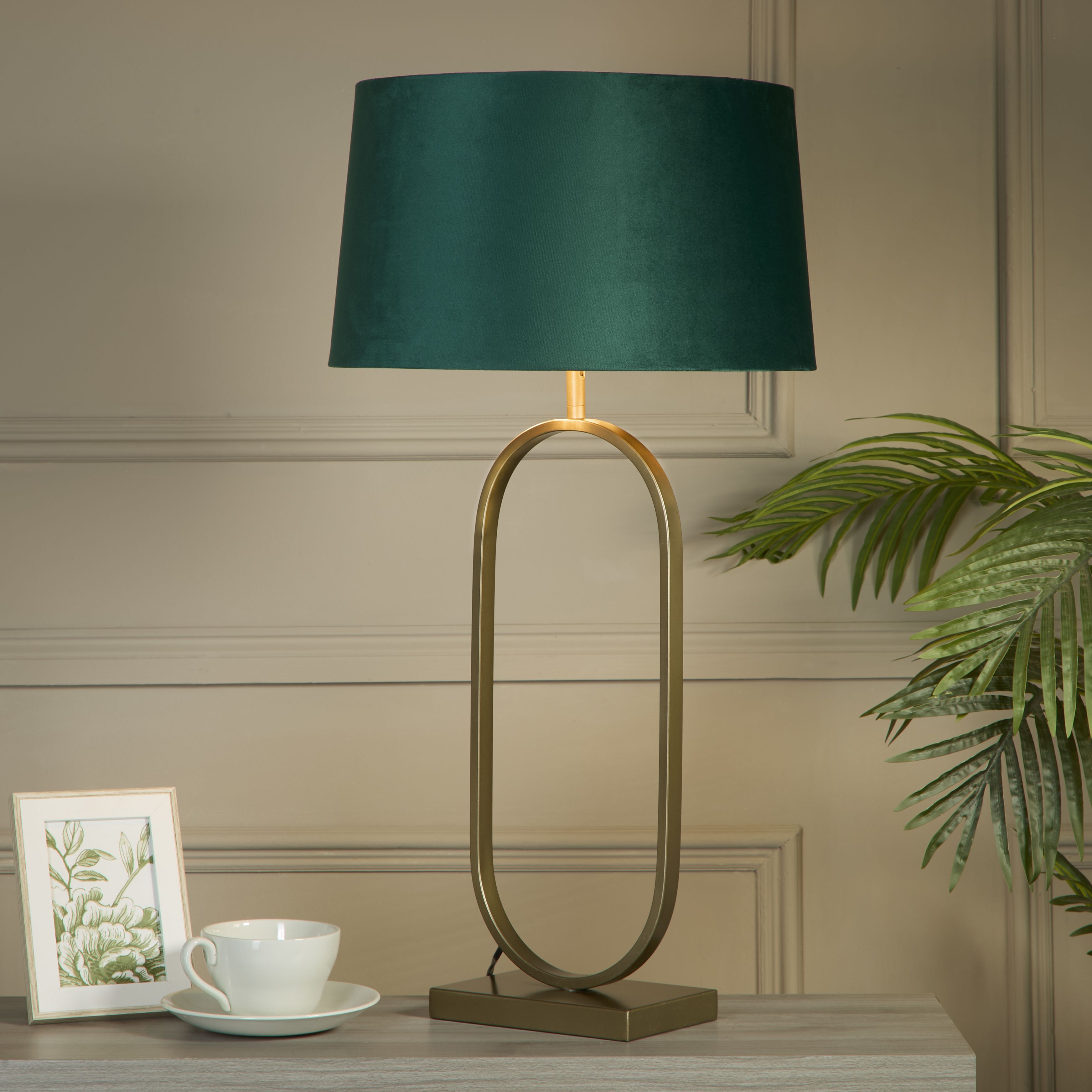 Bronze table lamp with an emerald velvet shade styled on a console table beside a teacup and saucer, a framed botanical print and a trailing palm plant, against a grey panelled wall