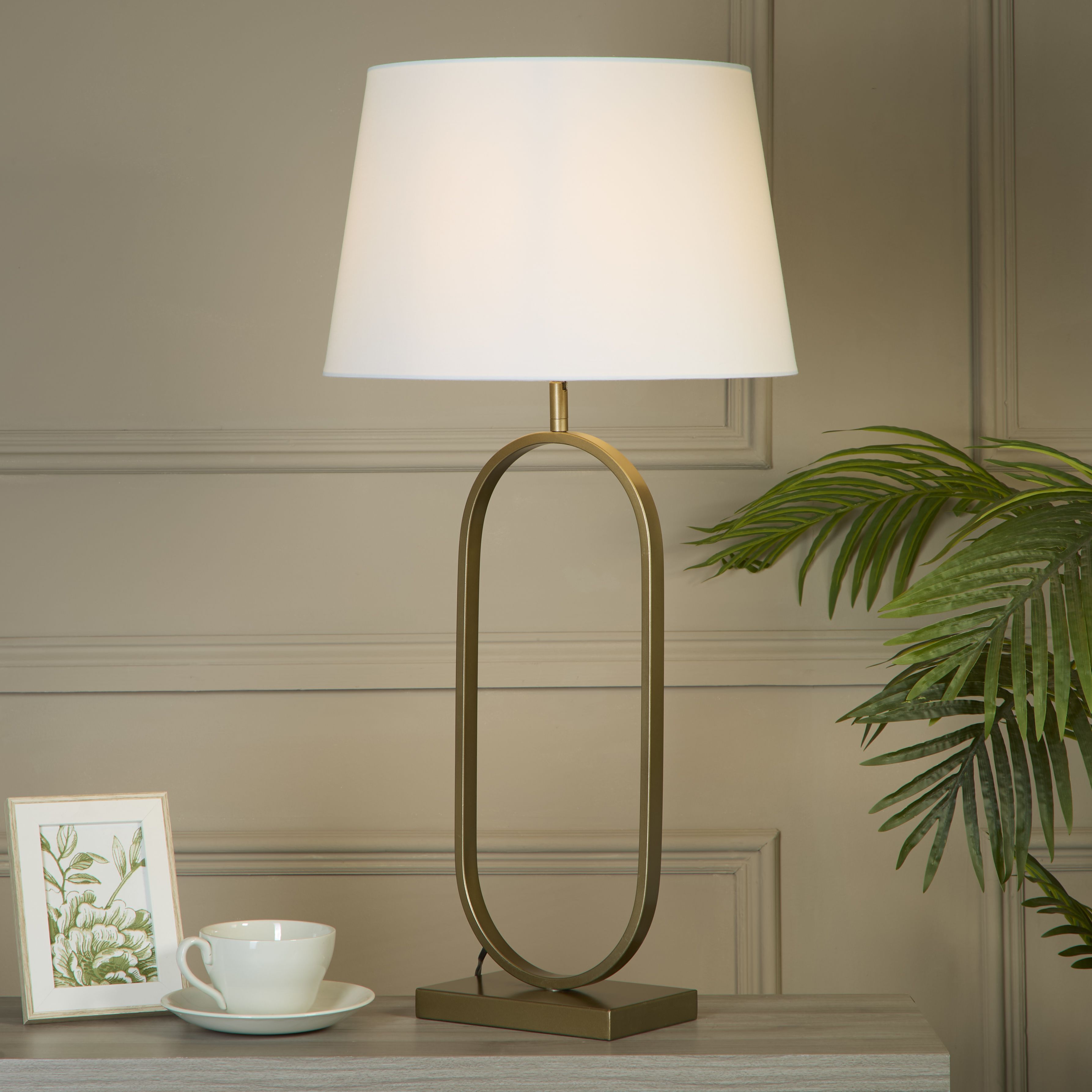 Bronze table lamp with a crisp white cotton shade styled on a console table beside a teacup and saucer, a framed botanical print and a trailing palm plant, against a grey panelled wall