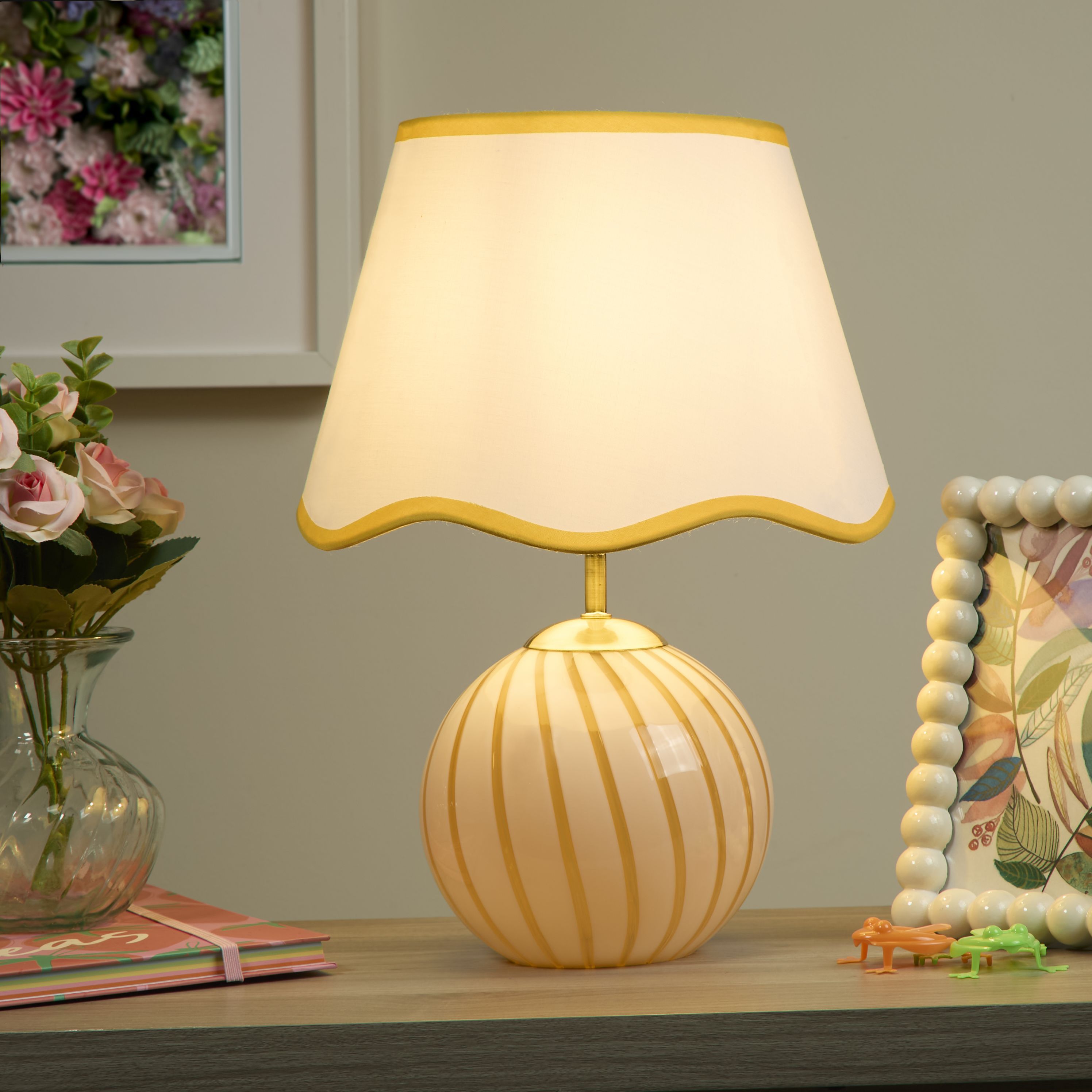 Celine yellow striped table lamp styled on a shelf with flowers, a picture frame and small decorative toys, lit with a warm glow