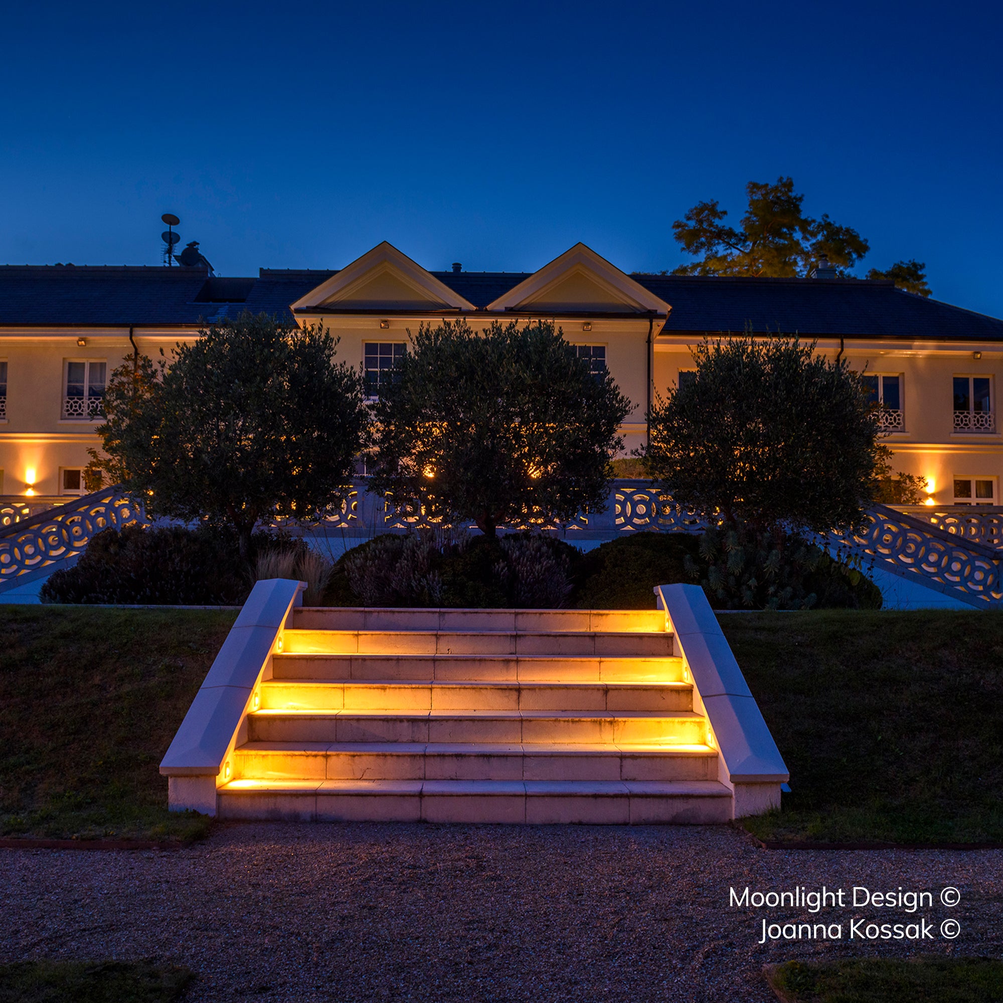 A wide symmetrical stone staircase leading up to a grand house, lit by recessed louvred step lights washing light evenly across each step at dusk. A Moonlight Design installation, not stock imagery.