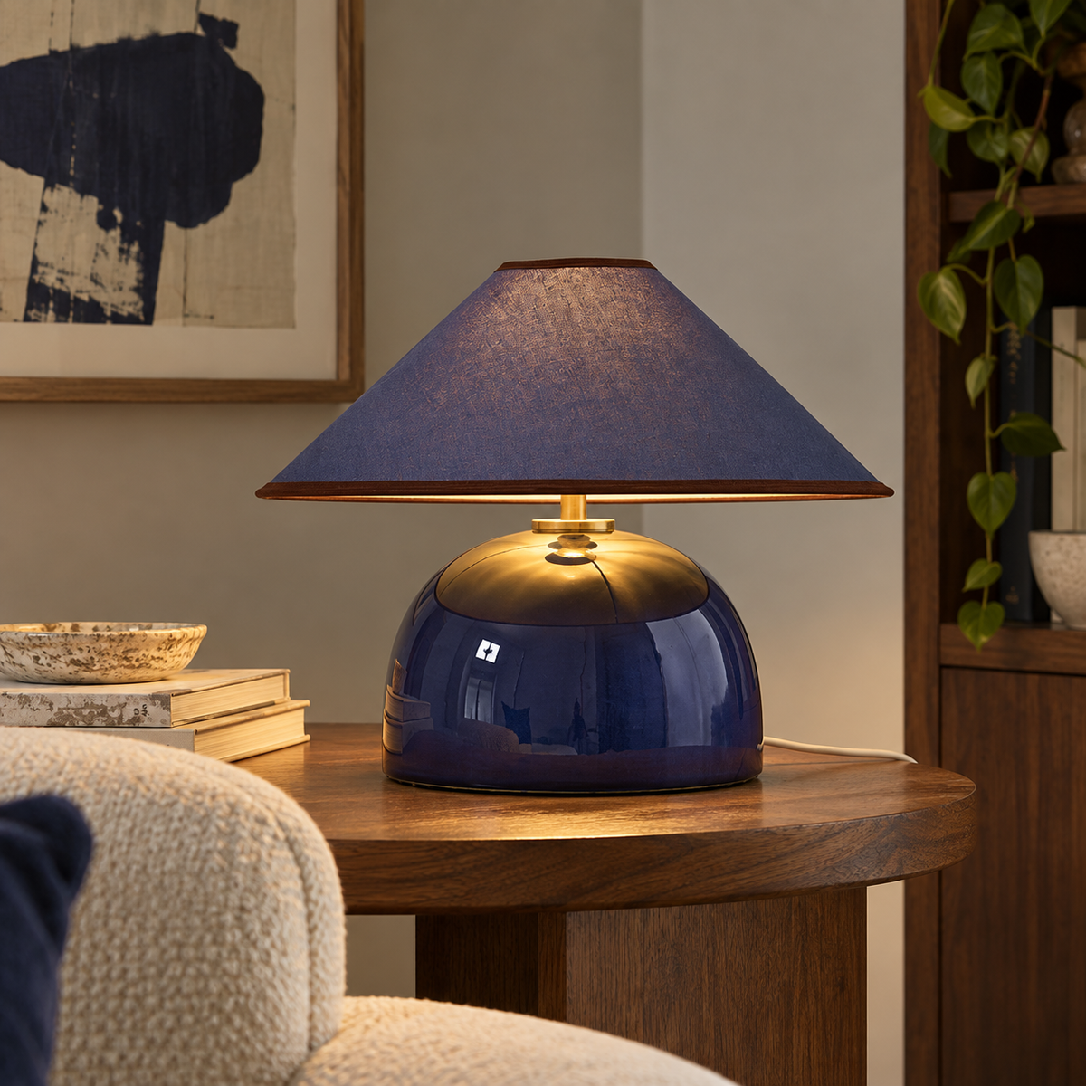 Nora navy blue ceramic table lamp with matching linen shade and antique brass details, lit on a wooden side table.