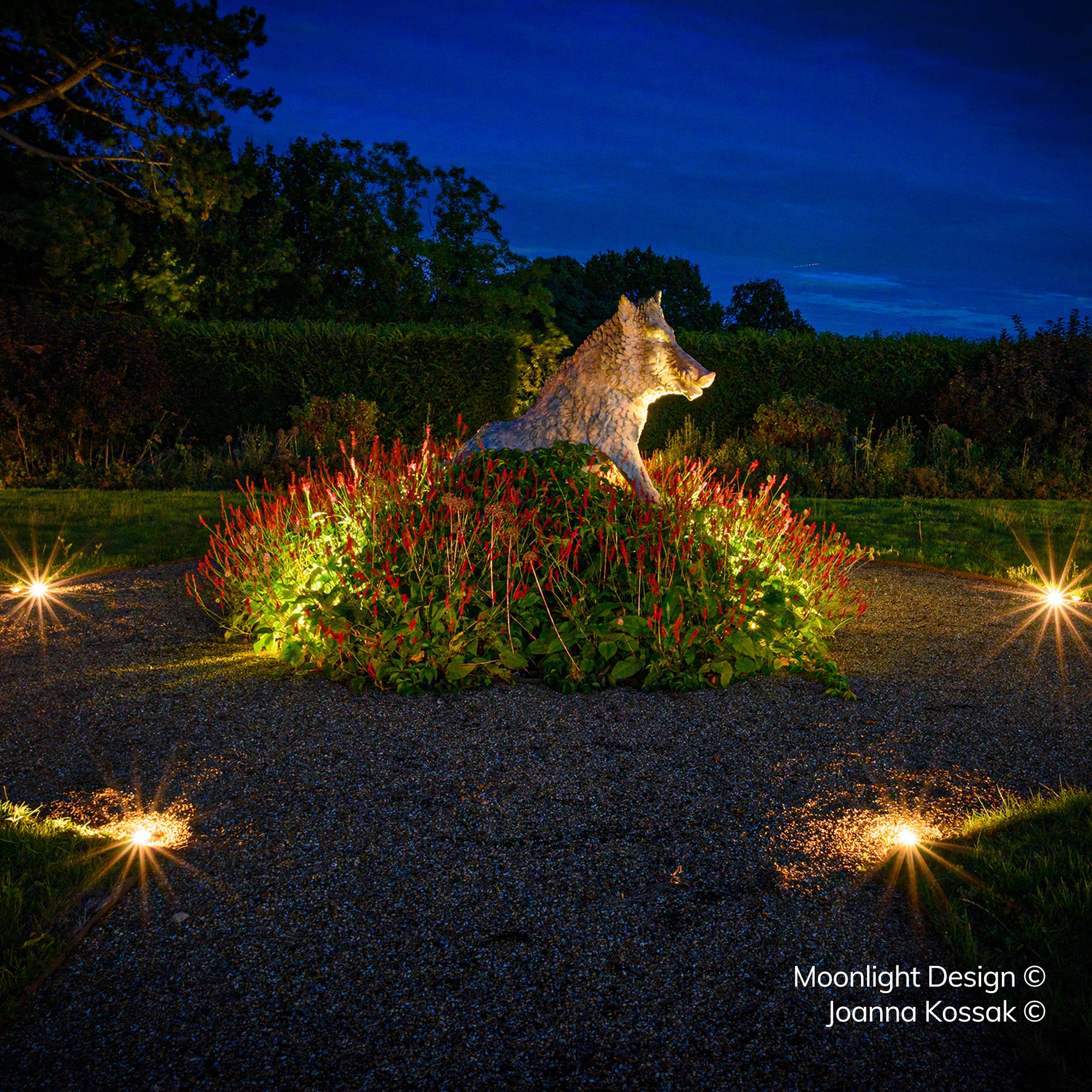 A stone boar sculpture in a floral bed at the centre of a gravel driveway loop, edged by multiple recessed Hunza Path Lights creating starburst points of light at night. A Moonlight Design installation, not stock imagery.