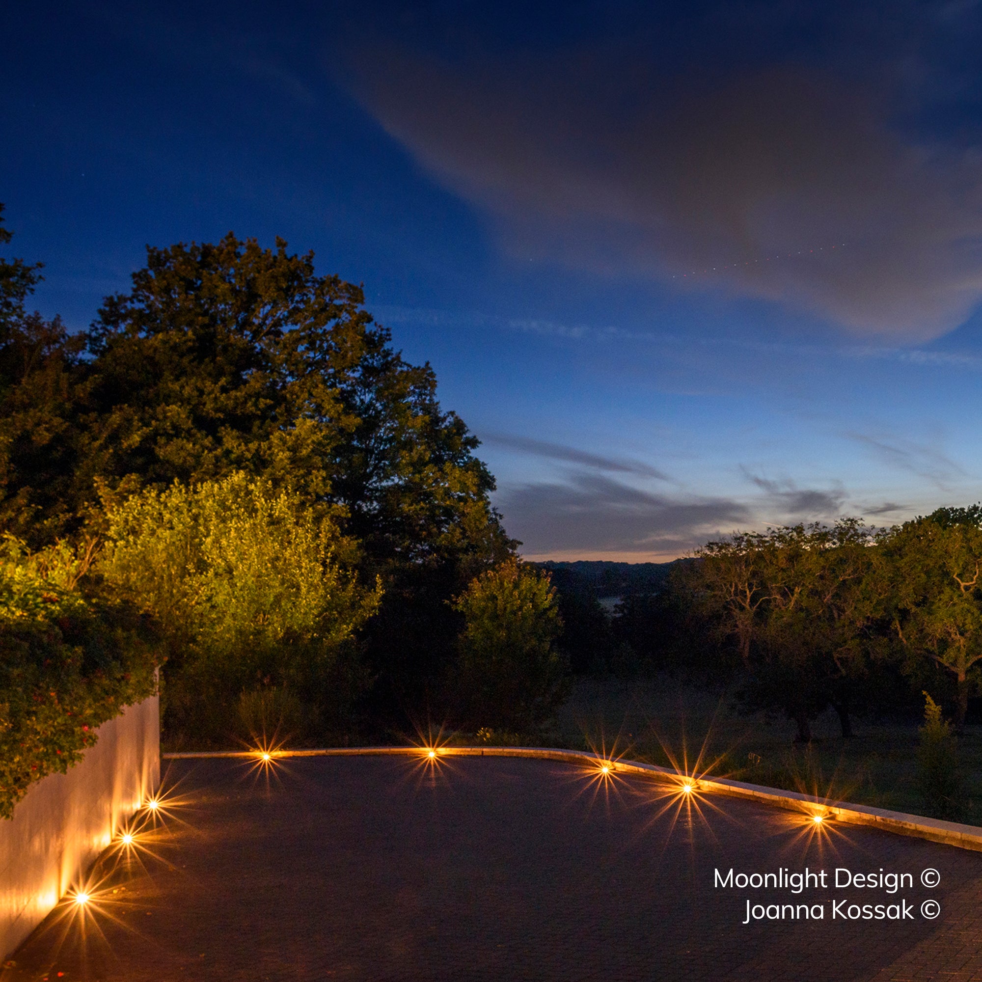 A curved gravel driveway at dusk, its edge lit by a run of recessed Hunza Path Lights along the kerb, with countryside visible beyond. A Moonlight Design installation, not stock imagery.