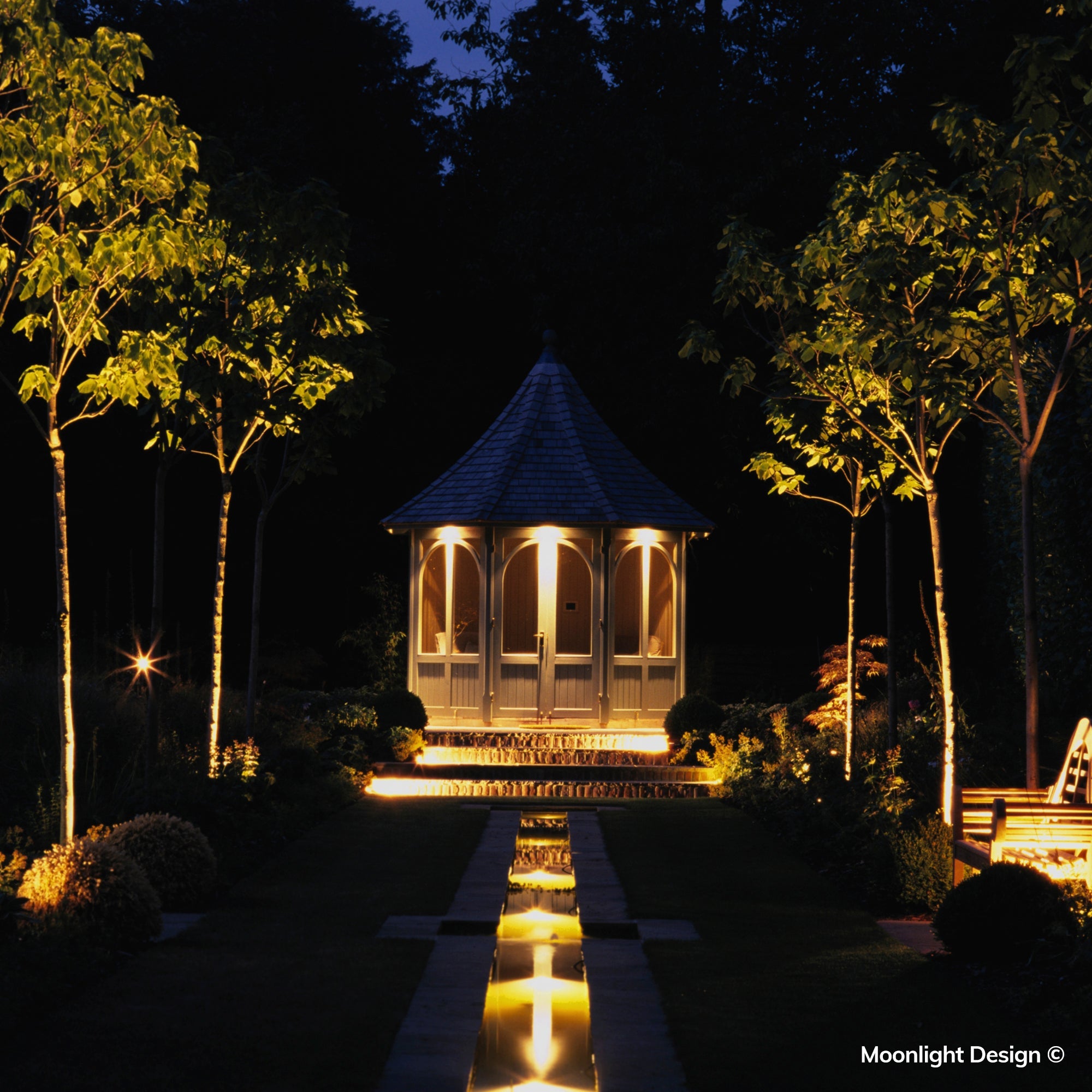 Hunza pond lights create a glowing reflection through the central water feature, drawing the eye towards the garden room and illuminated trees beyond. A completed Moonlight Design pond lighting project.