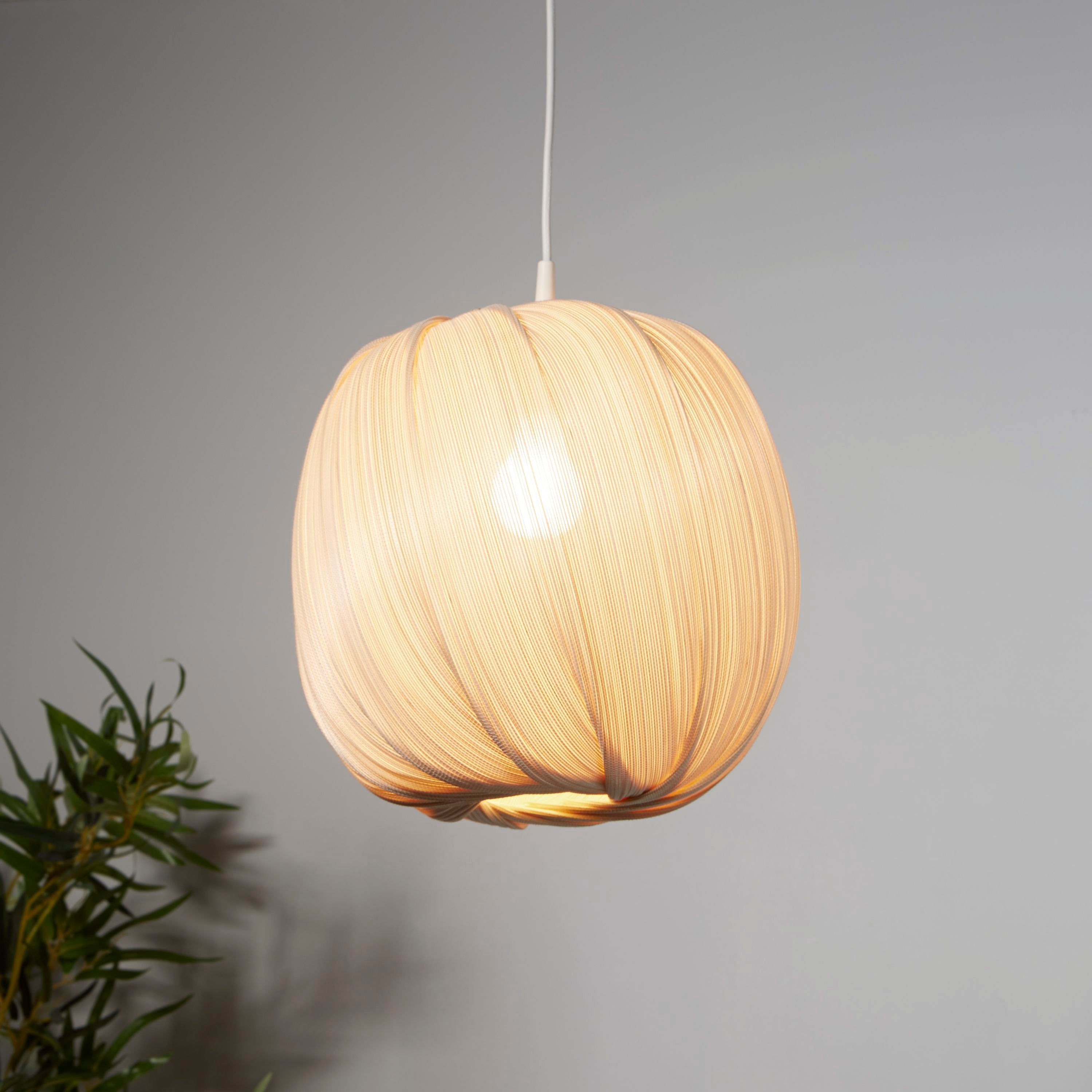 Searchlight natural round bamboo shade lit and hanging against a grey wall, styled with a green plant in the foreground