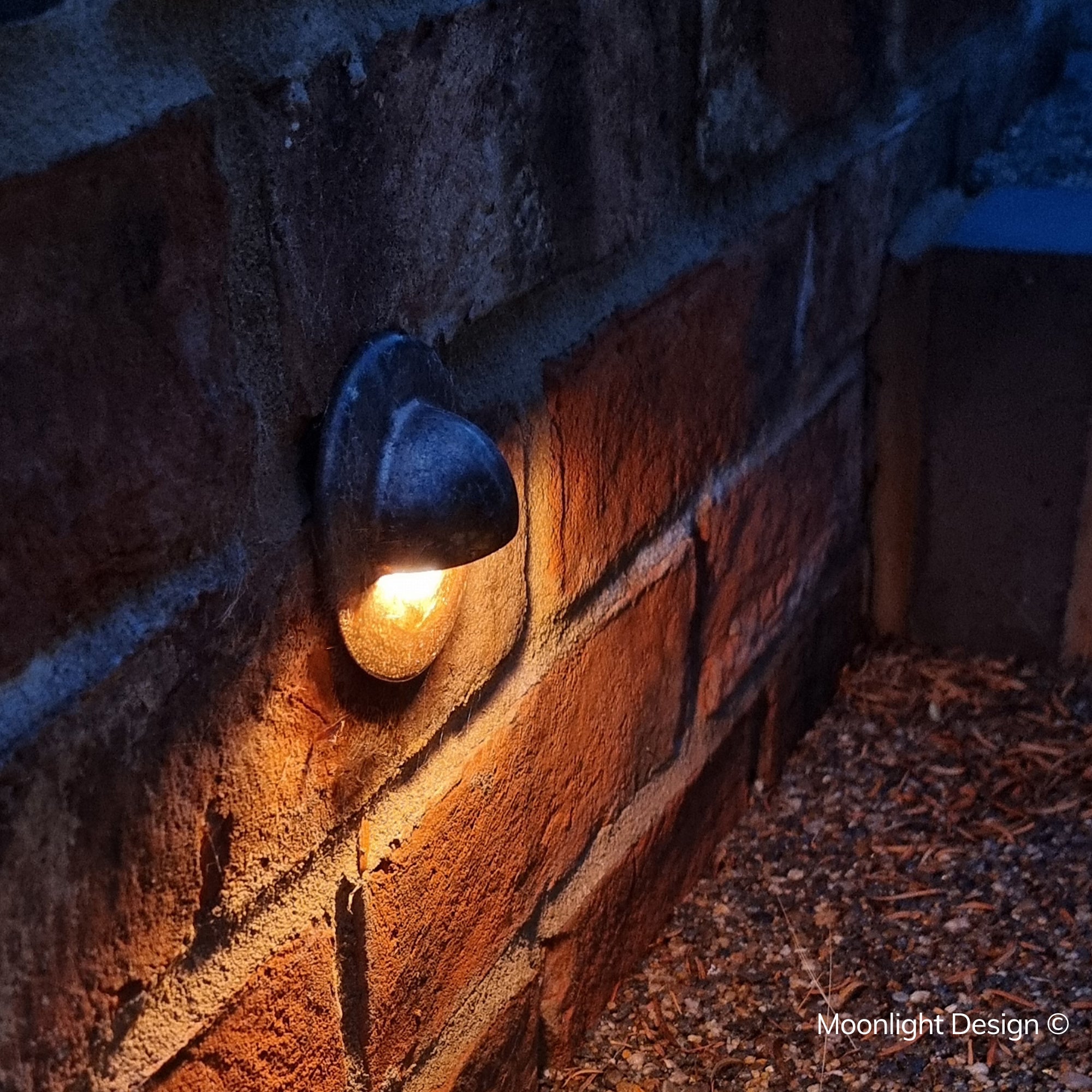 Hunza eyelid step light recessed into a rendered stone wall base, casting light across brick paving at night. A Moonlight Design installation, not stock imagery.