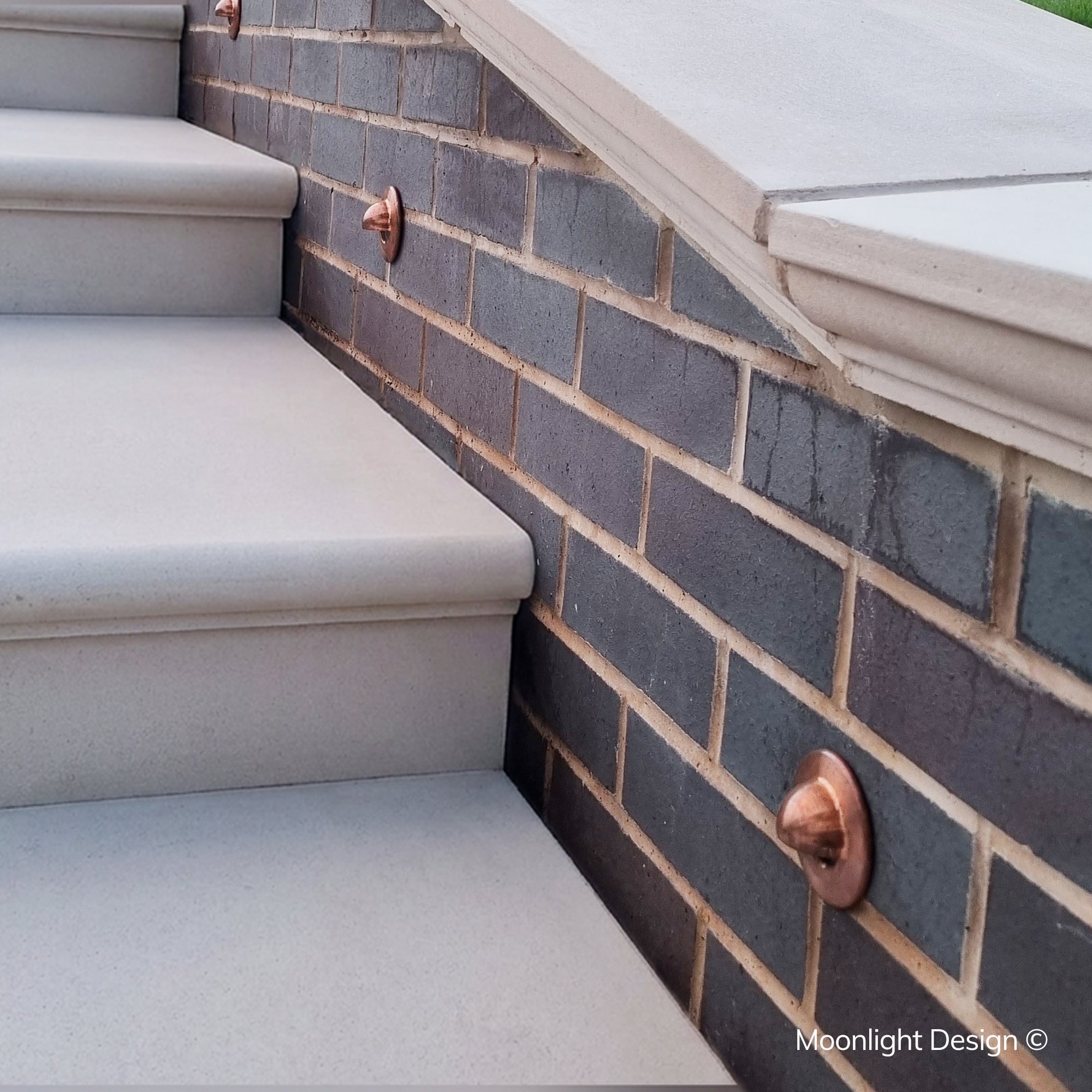 Two Hunza eyelid step lights in copper recessed into dark grey brick step risers, lighting the tread of each step at night. A Moonlight Design installation, not stock imagery.