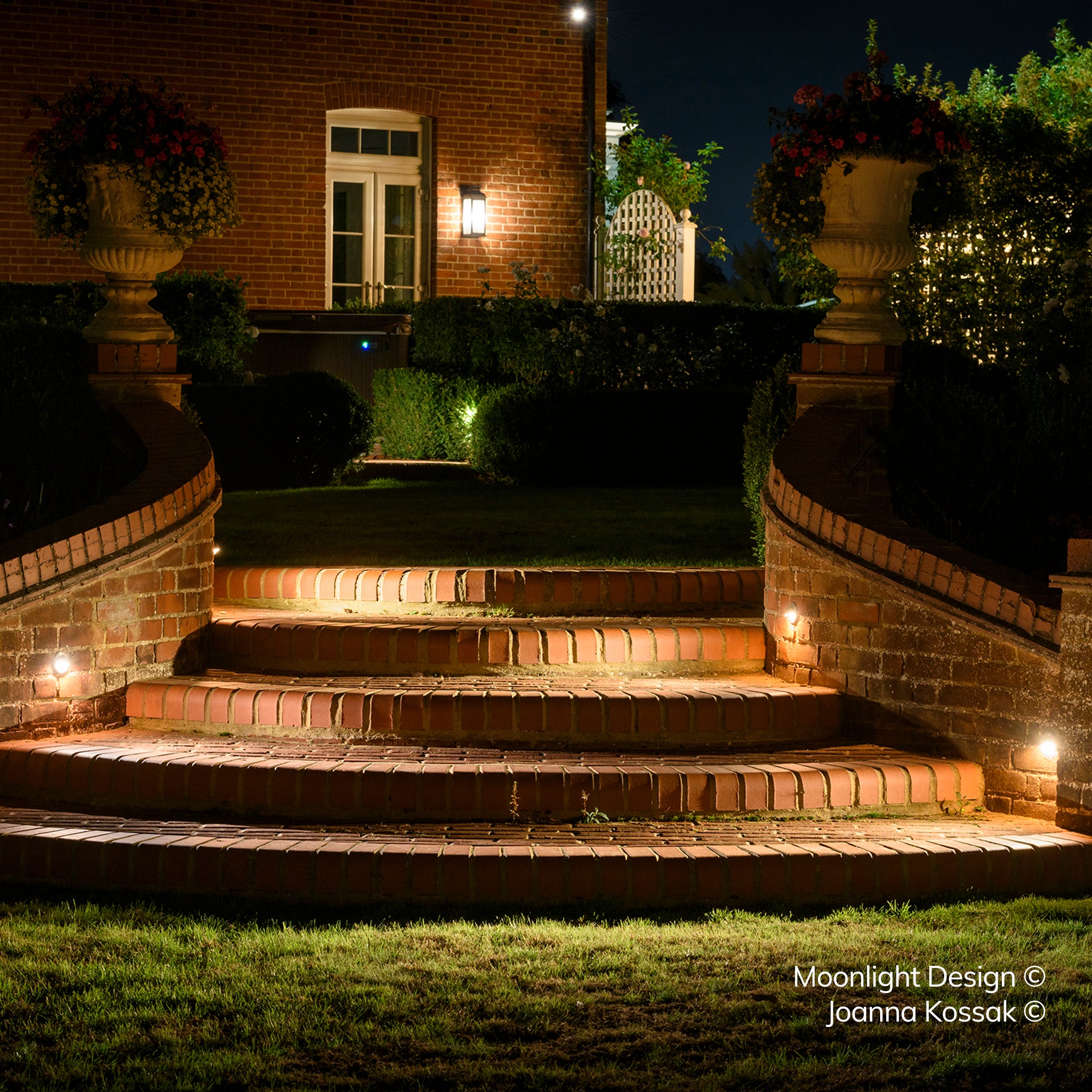 Curved brick garden steps leading up to a house, lit by recessed Hunza eyelid step lights along the risers and low brick walls either side, with planted stone urns above. A Moonlight Design installation, not stock imagery.