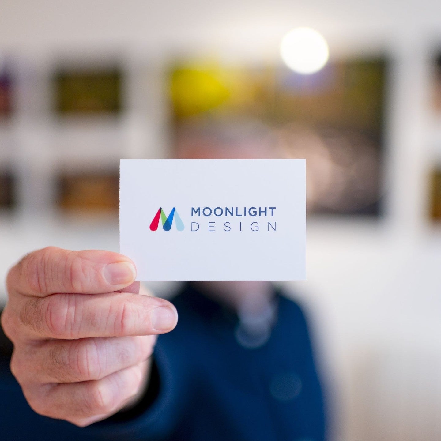 Hand holding a business card with 'Moonlight Design' logo against a blurred indoor background