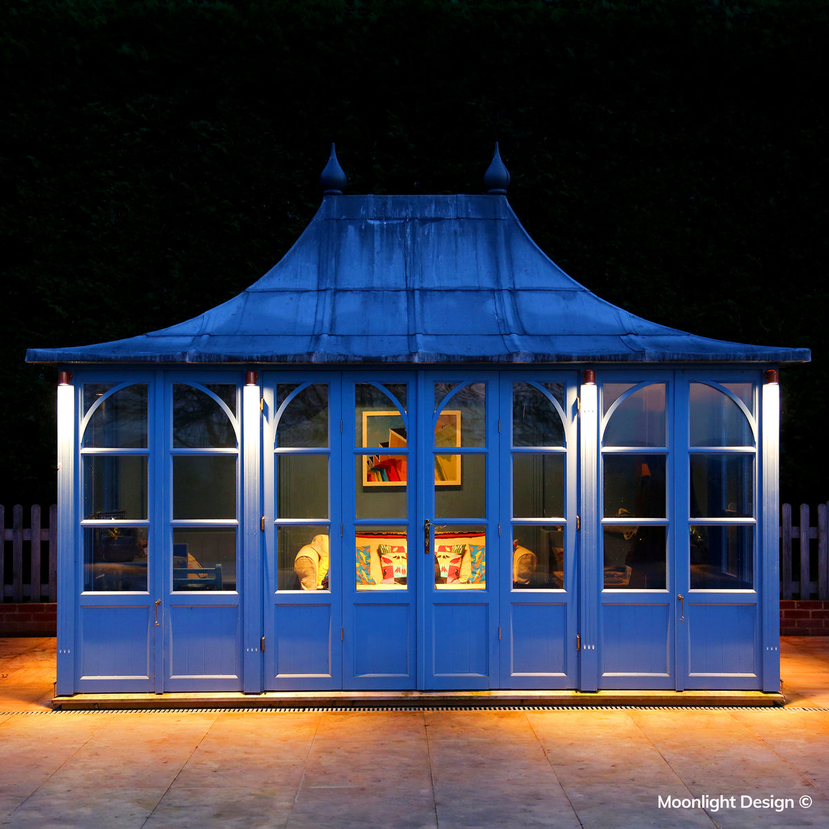 Moonlight Design lifestyle photograph of Hunza wall down lights washing light down the corner posts of a blue-painted garden pavilion at dusk.