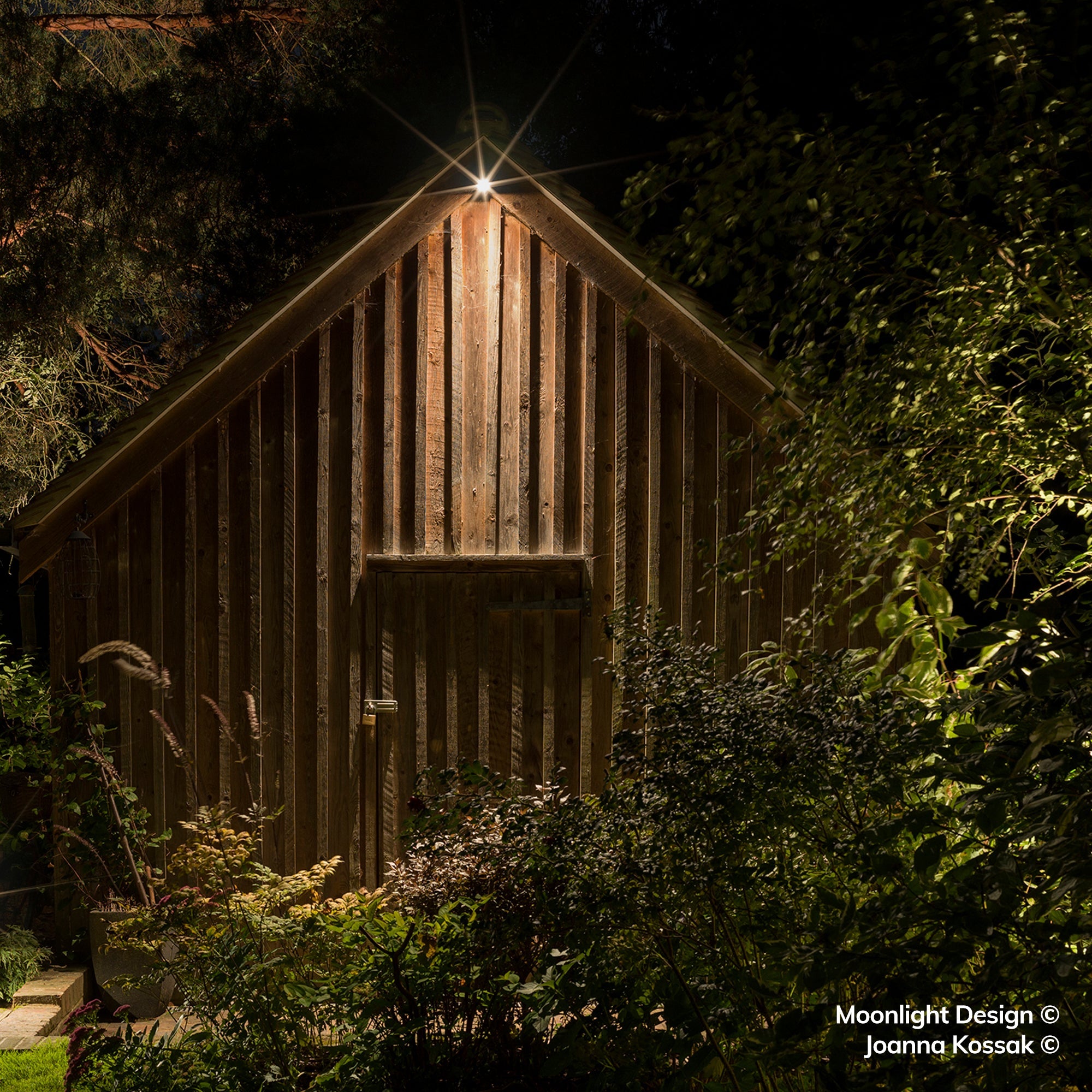 Moonlight Design lifestyle photograph of a Hunza wall down light at the peak of a timber gable, illuminating the vertical cladding and door of a garden building at night.