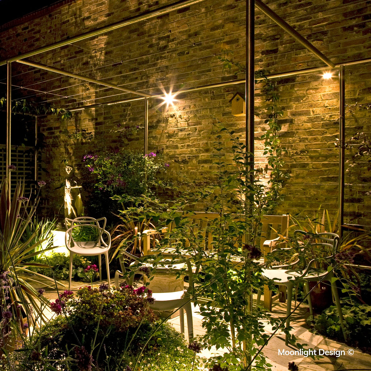 Moonlight Design lifestyle photograph of Hunza wall spots lighting a brick garden wall and birdhouse within a pergola seating area, with a garden statue and flowering plants.