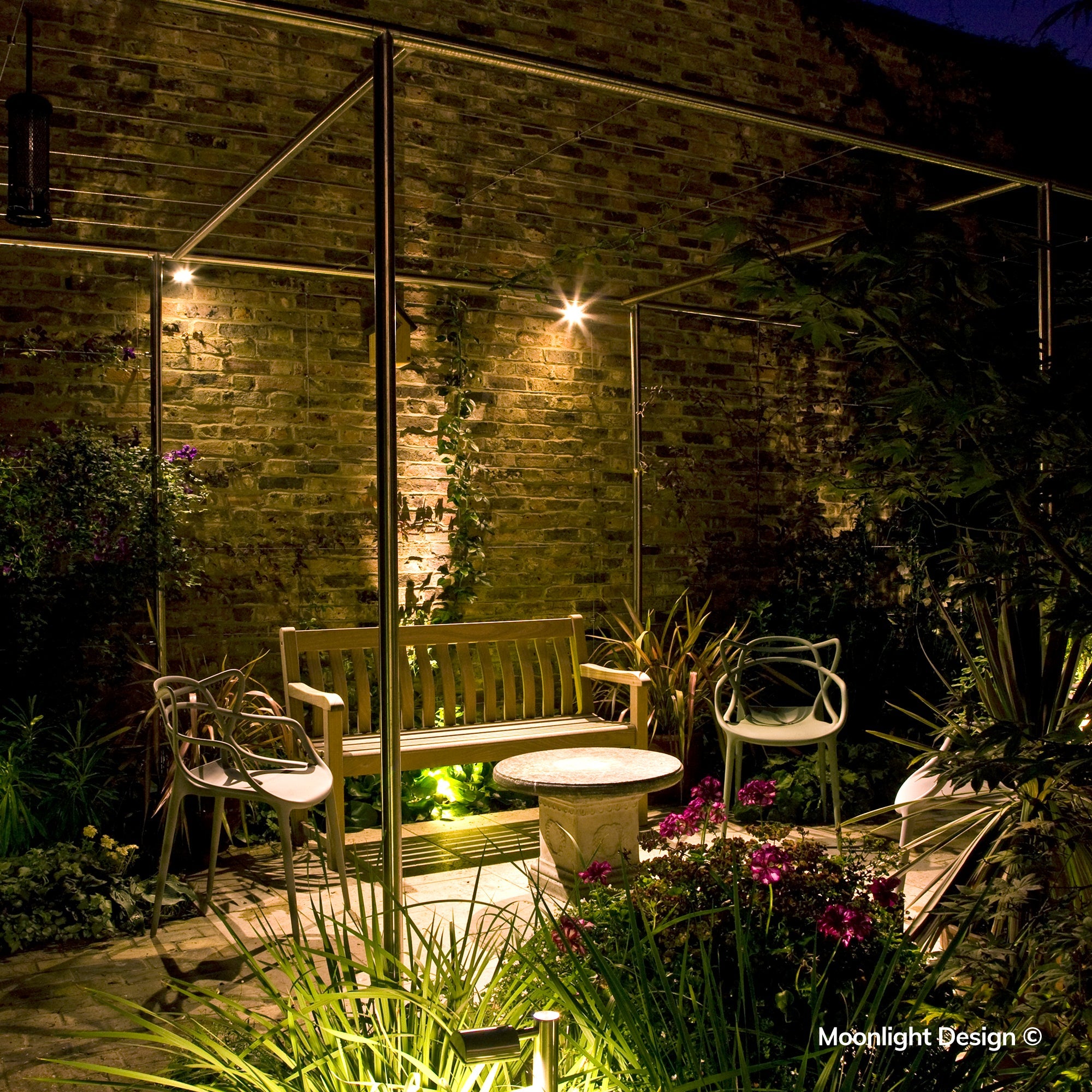 Moonlight Design lifestyle photograph of a Hunza wall spot lighting a brick wall and birdhouse above a wooden bench in a pergola-covered garden seating area at night.