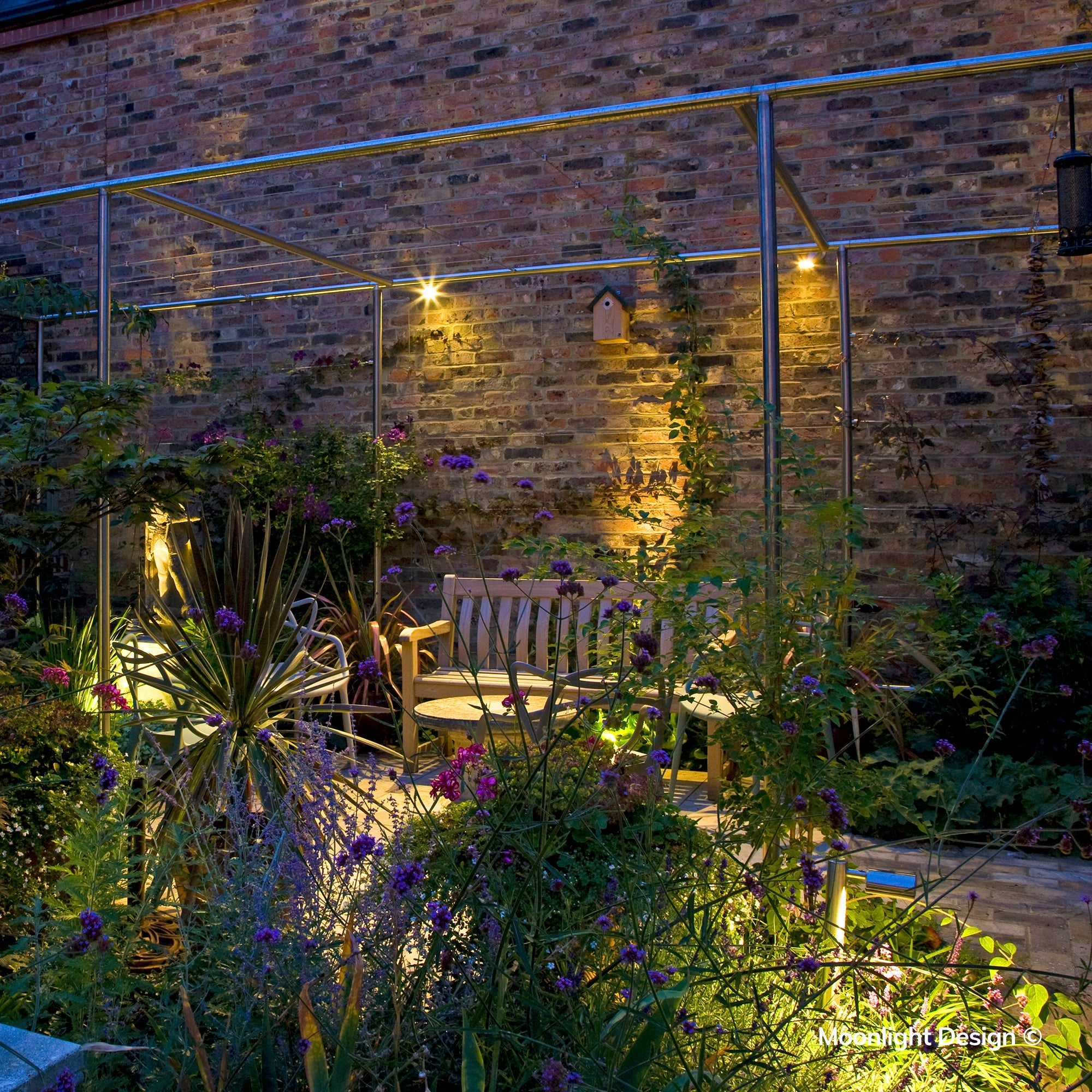 Moonlight Design lifestyle photograph of a Hunza wall spot lighting a brick wall and birdhouse above a garden bench and table, in a pergola seating area with flowering plants at night.
